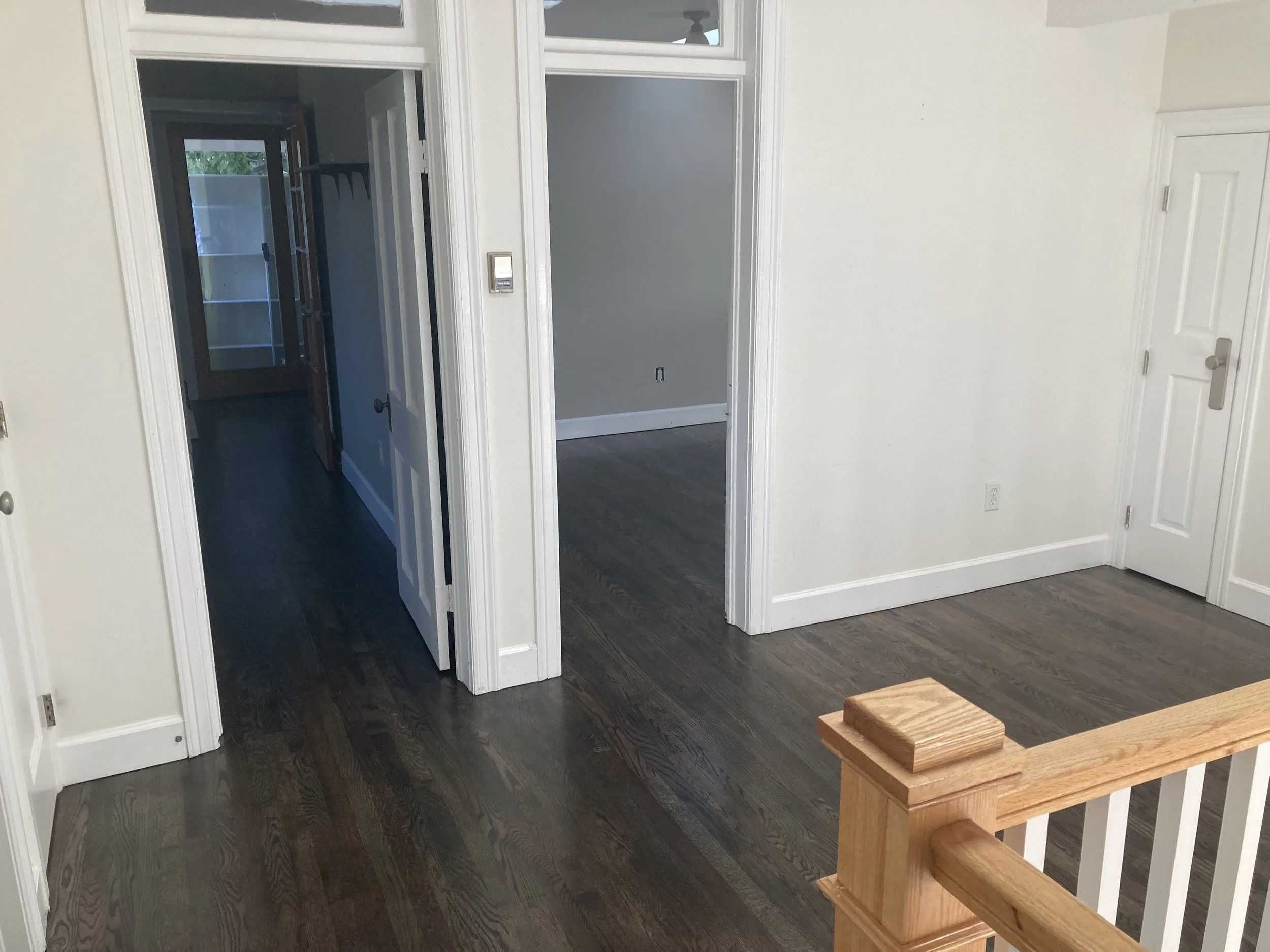 Interior view of a house with hardwood flooring, white walls, white doors, and doorways leading to other rooms. Part of a wooden staircase railing is visible in the foreground.