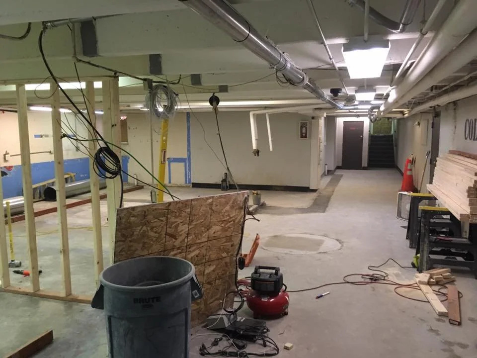 Construction site indoors with exposed pipes, electrical wiring, and partially built walls. There are tools, a trash can, and lumber stacks visible.
