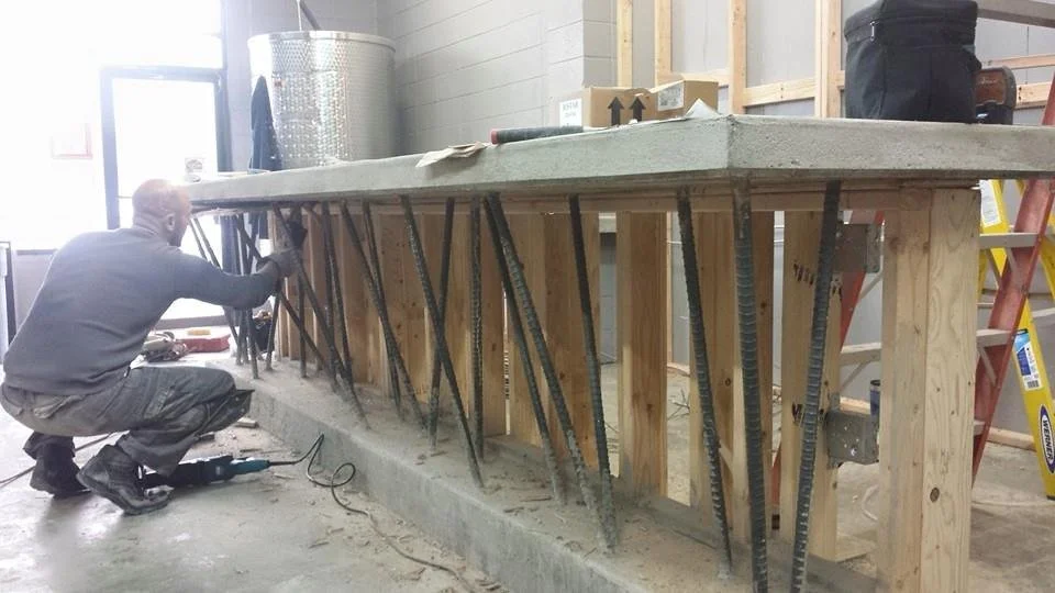 A worker crouched down working on a construction project, installing or securing steel rebar supports beneath a concrete countertop or slab, in an indoor construction site with tools and materials around.