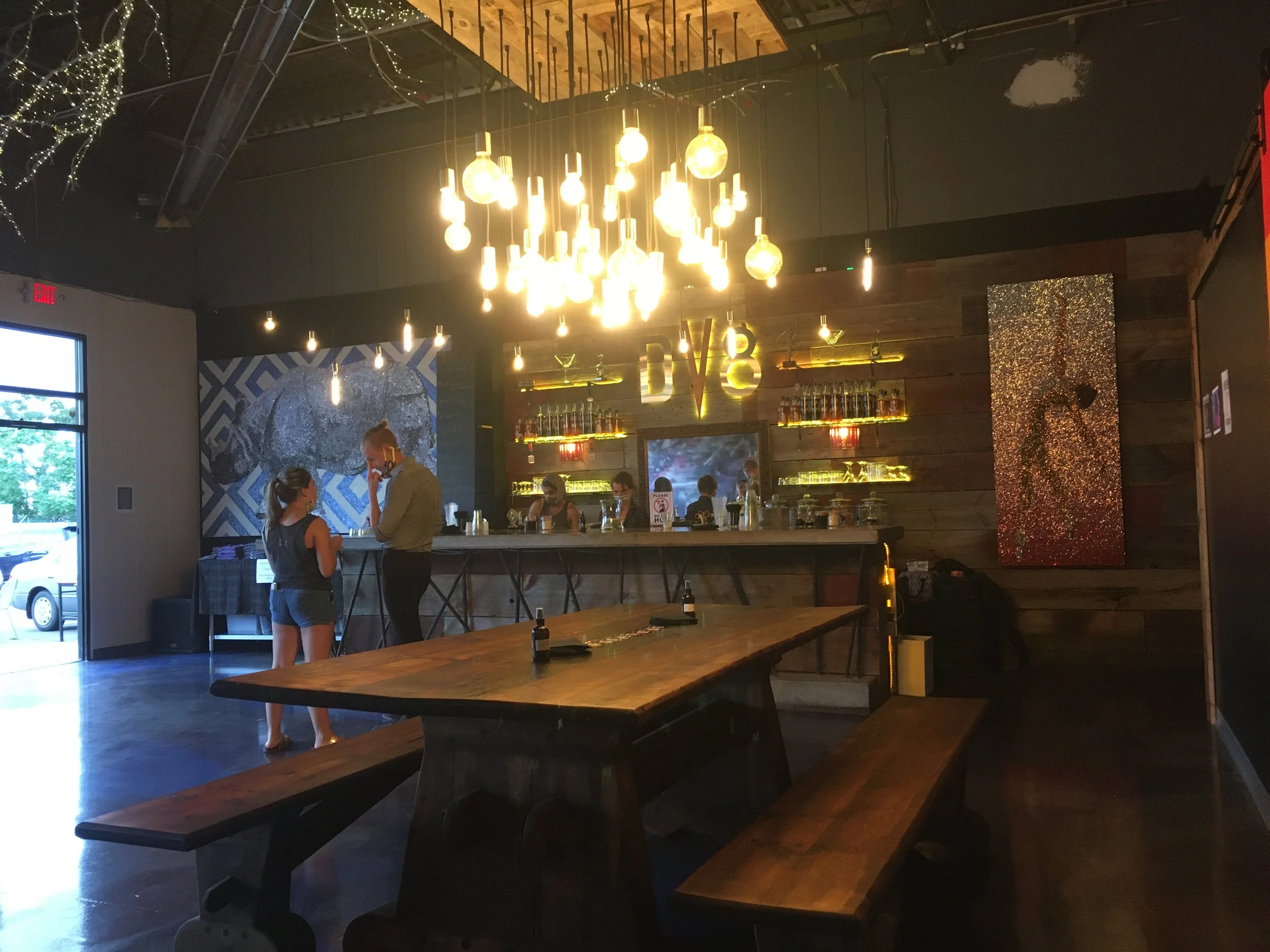 Inside a dimly lit bar with pendant lights hanging from the ceiling, a wooden wall behind the bar features a neon sign that says 'LOVE'. There is a large wooden communal table with benches in the foreground. Two women are near the bar, talking, with other people sitting at the bar in the background. Artwork and bottles of alcohol are visible behind the bar.