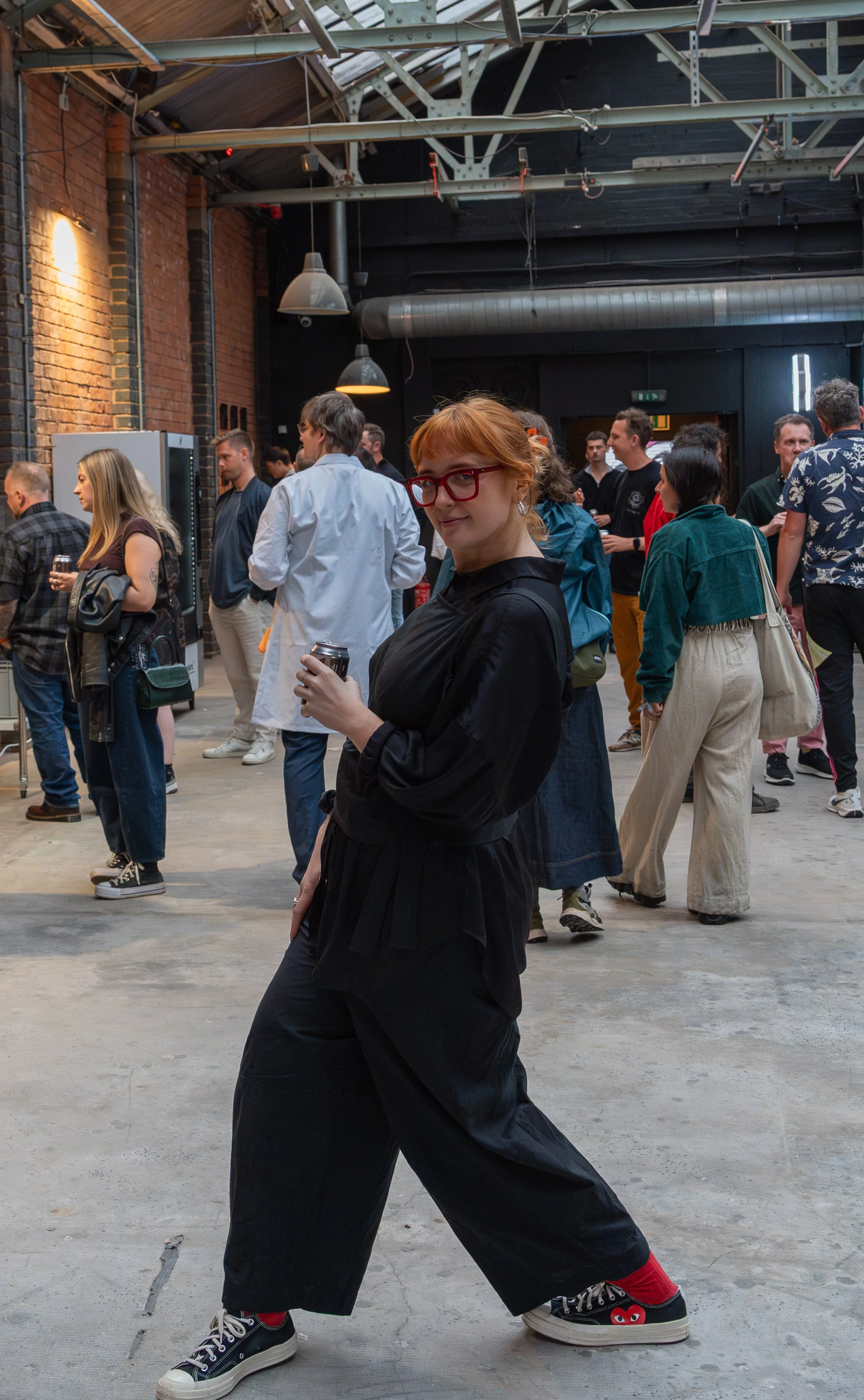 A woman with red hair and red glasses standing in a warehouse or industrial space at a social gathering, holding a drink and smiling at the camera.