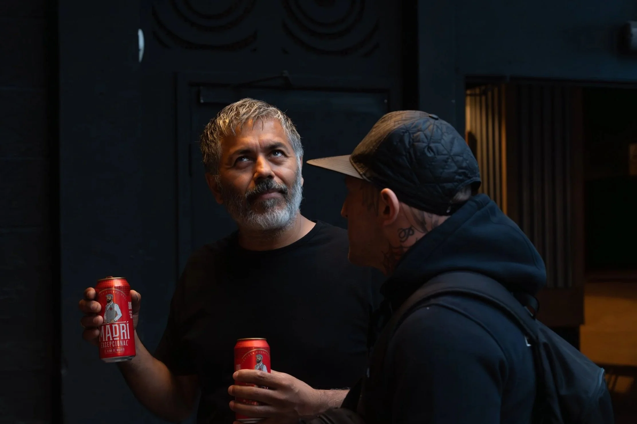 Two men having a conversation, one older with gray hair and beard holding a can of Madrí beer, and one younger with tattoos, wearing a cap and backpack.