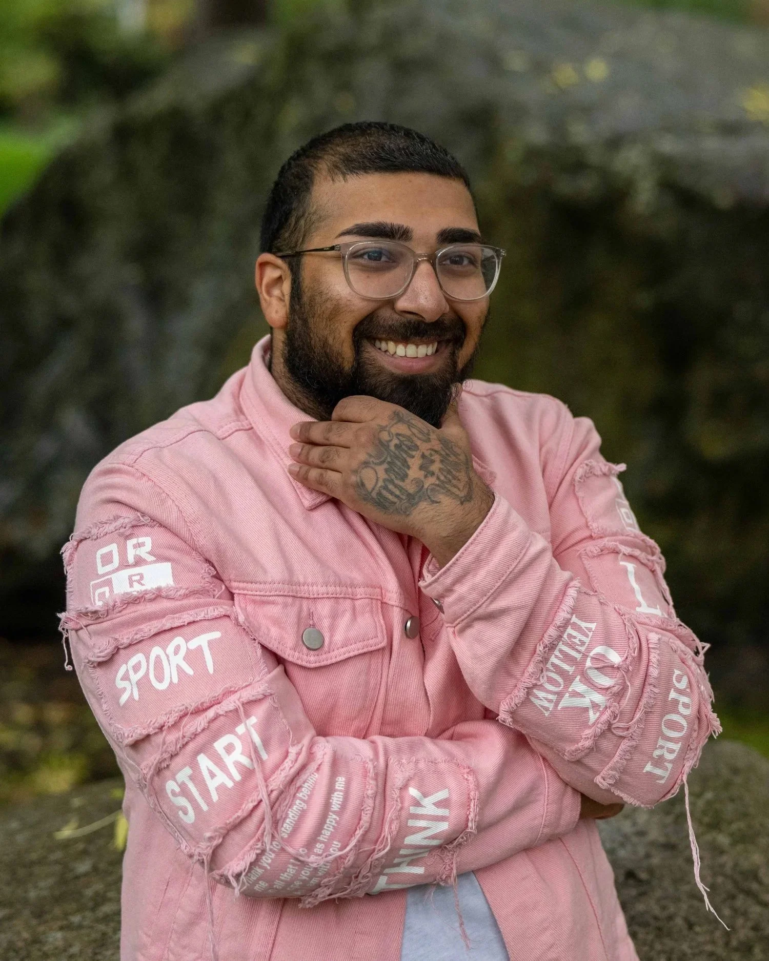 A smiling man with glasses, a beard, and tattoos on his hand, wearing a distressed pink jacket with words like 'SPORT,' 'START,' and 'THANK' on the sleeves, standing outdoors with a blurred green background.