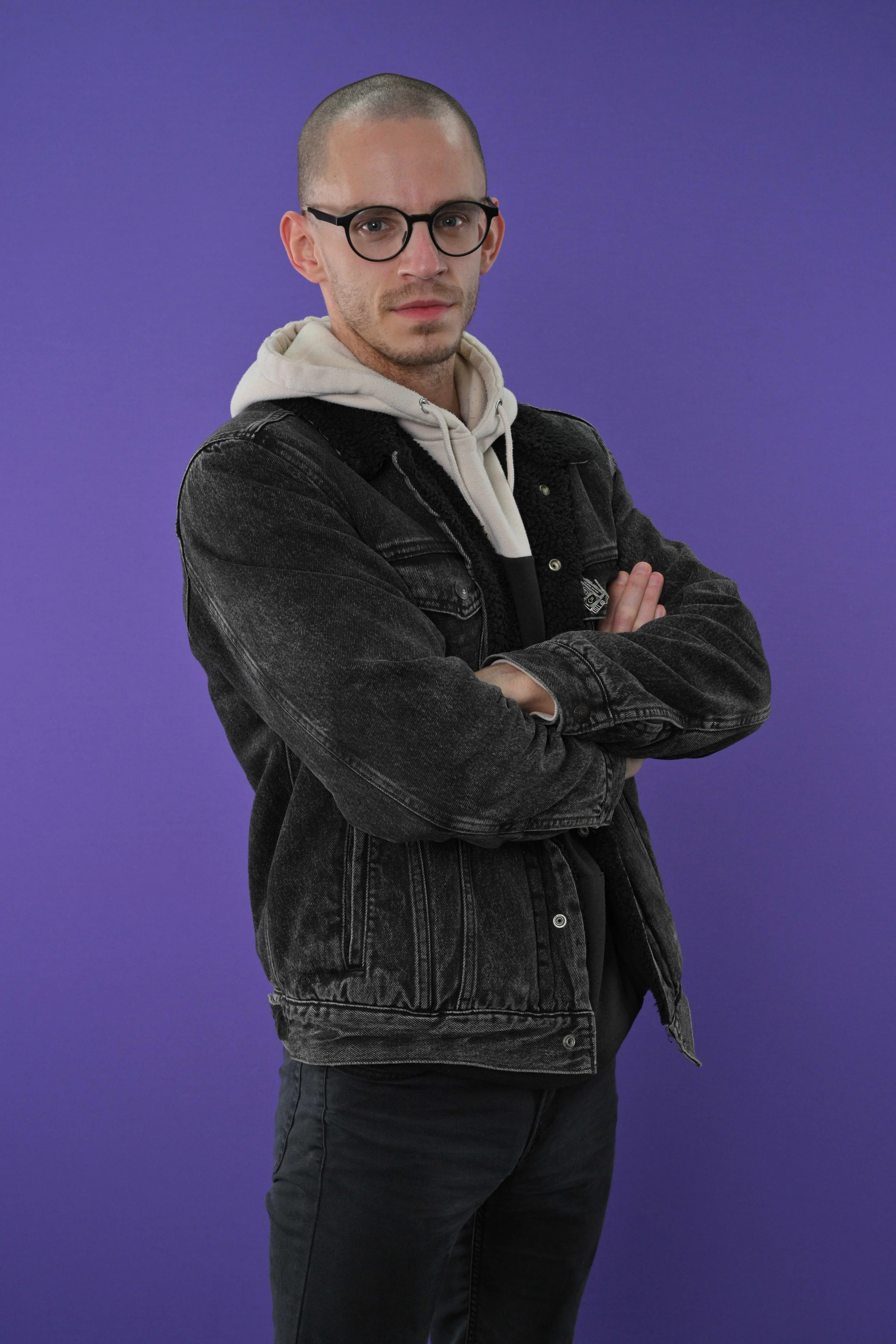 Young man with short hair and glasses, wearing a black denim jacket over a cream hoodie, standing against a purple background with arms crossed.