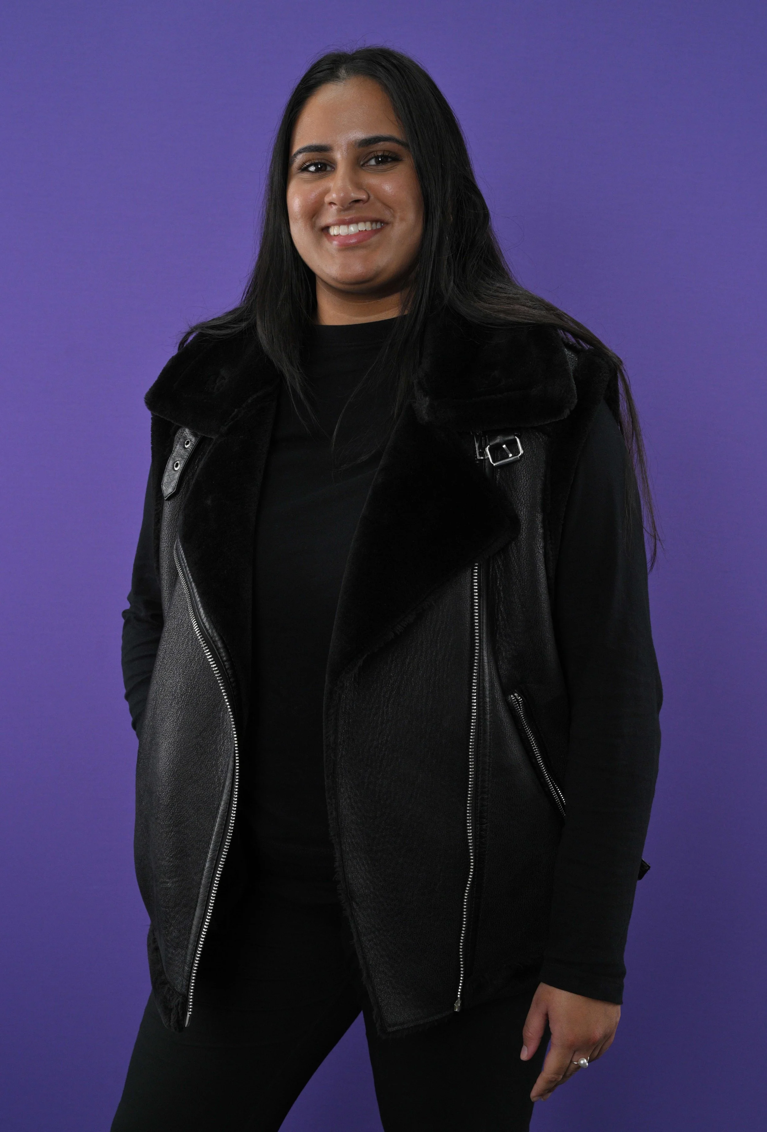 A woman with long black hair, wearing a black leather vest over a black top, standing against a purple background and smiling at the camera.