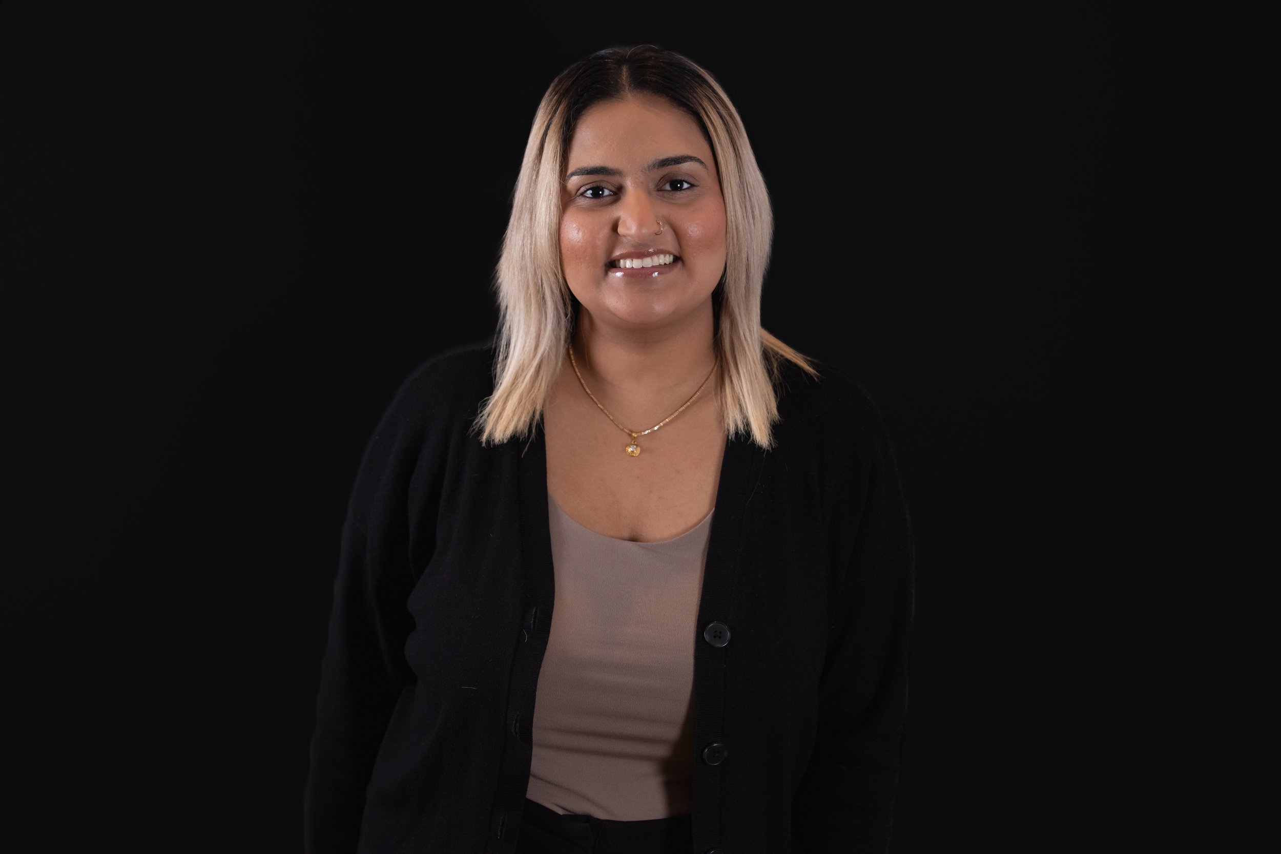 A portrait of a woman with shoulder-length blonde hair, smiling, wearing a black cardigan over a beige top and gold jewelry, against a black background.