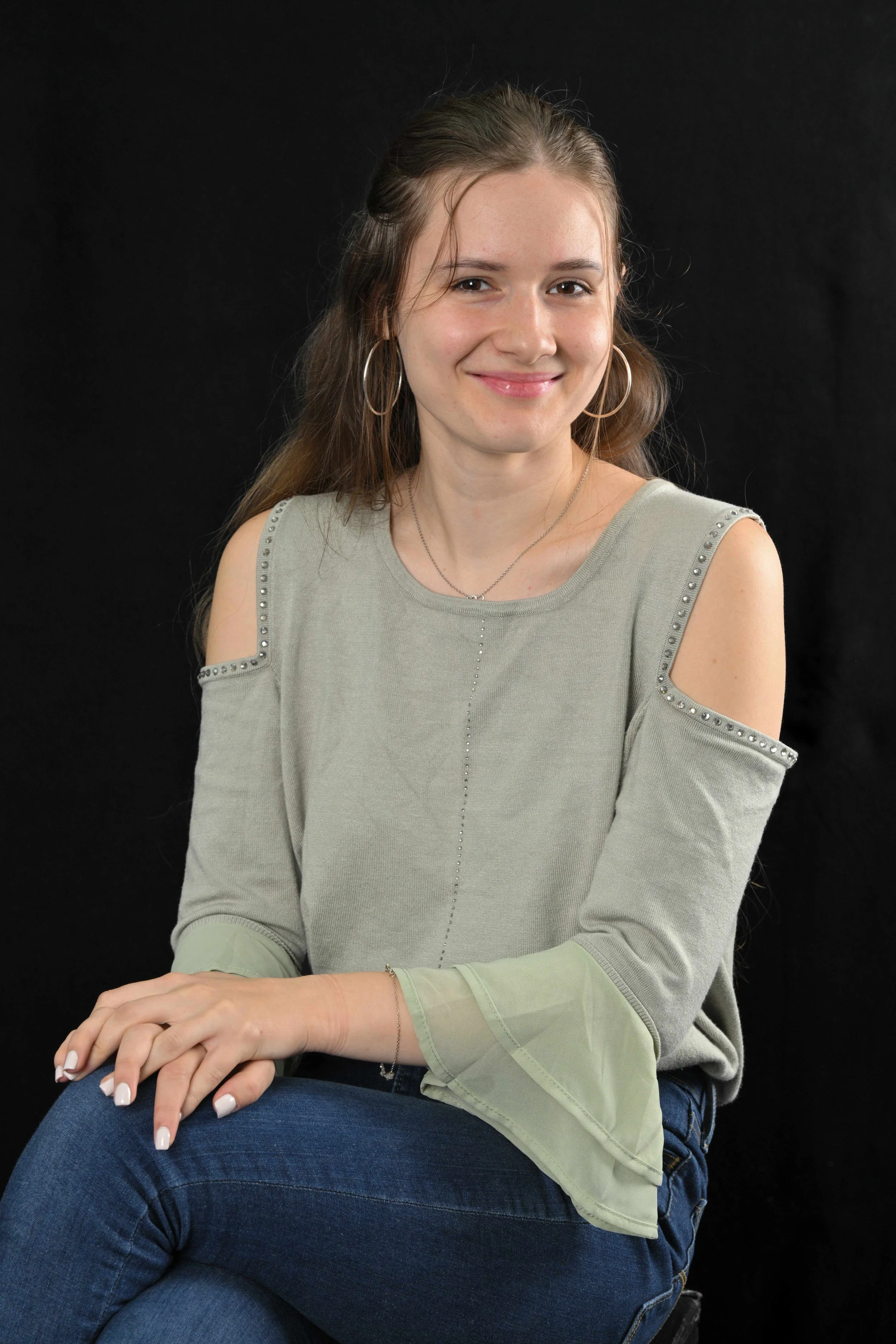 A young woman with long light brown hair, wearing hoop earrings, a layered necklace, and a light green cold-shoulder top with embellishments on the sleeves, sitting against a black background and smiling at the camera.