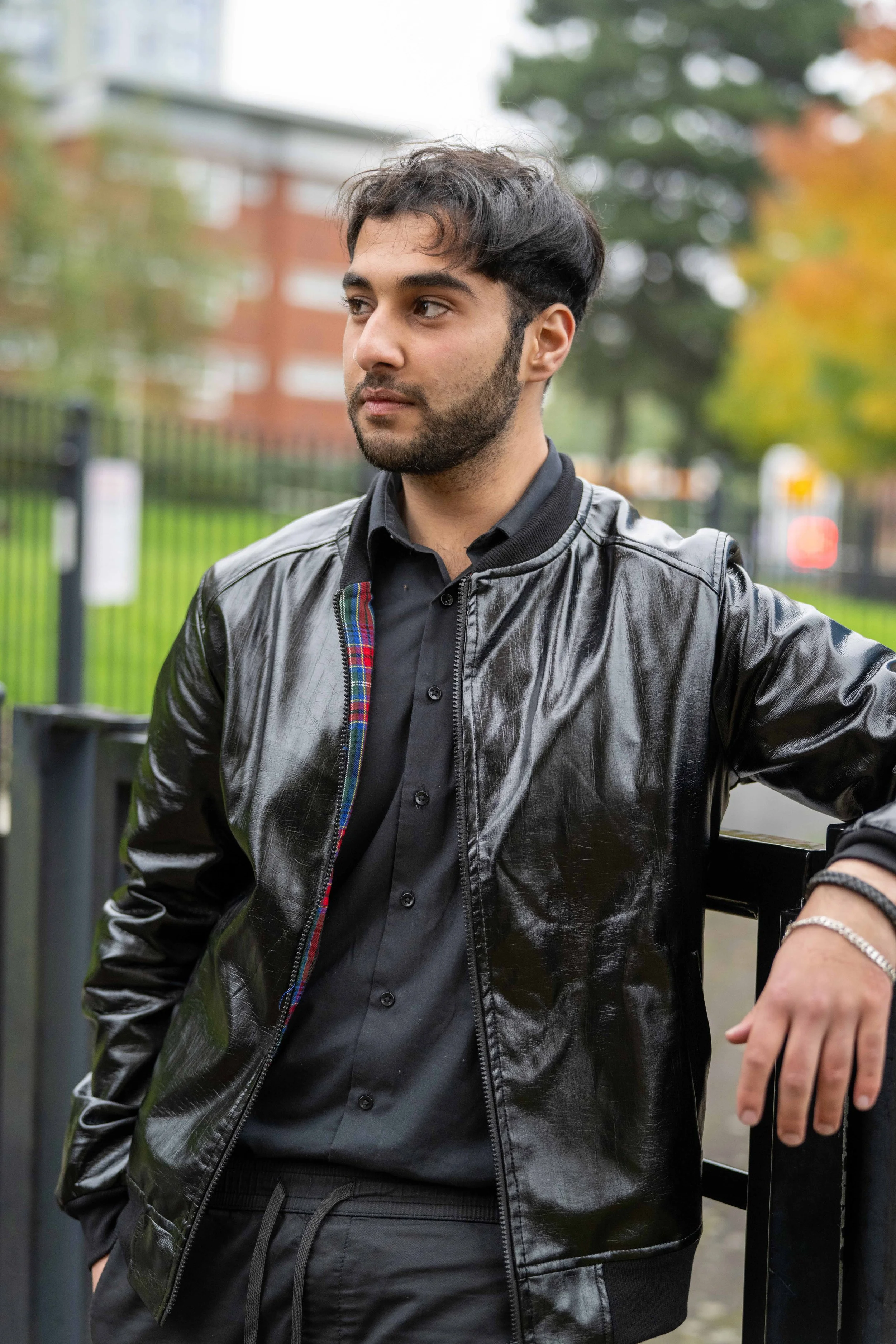 A young man with dark hair and a beard wearing a black leather jacket and black shirt, standing outdoors near a fence, with trees and a building in the background.