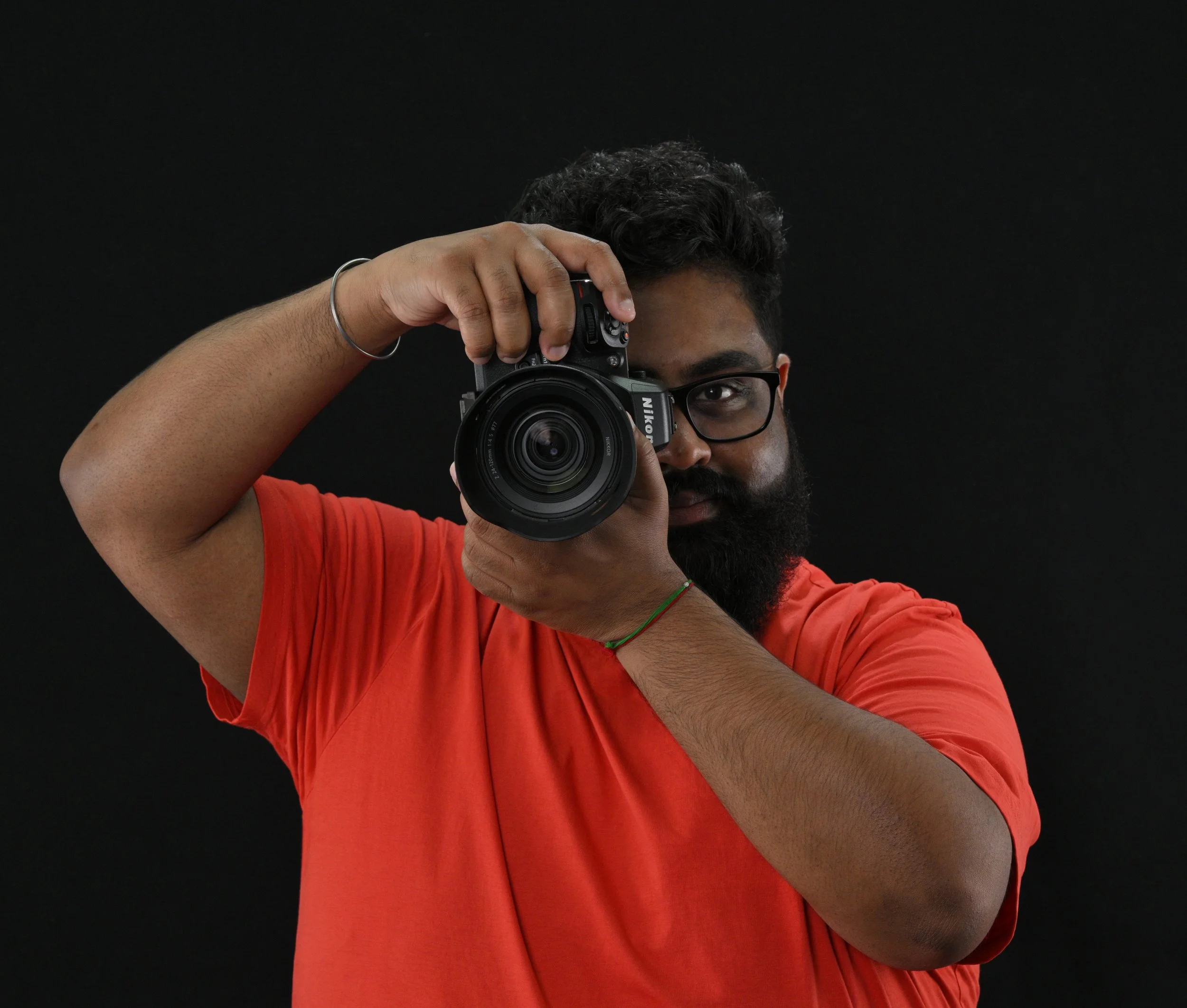 A man with dark hair, glasses, and a beard is holding a camera up to his eye, looking through the viewfinder while taking a photo. He is wearing a red shirt and standing against a black background.