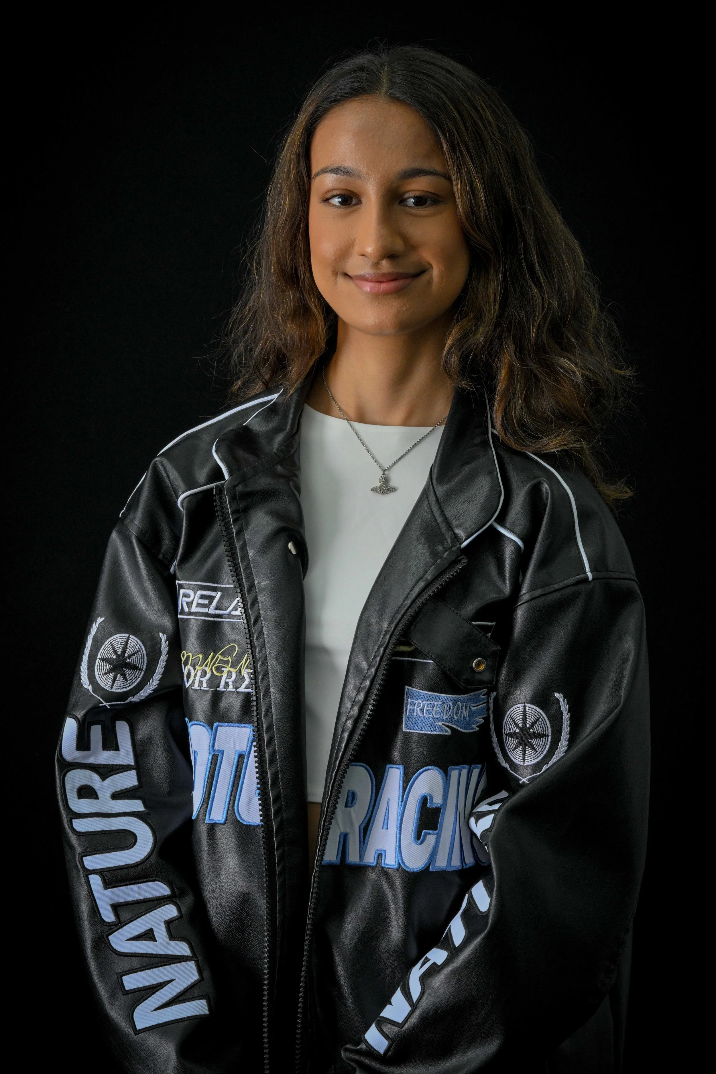A young woman with long, dark, wavy hair smiling, wearing a black racing jacket adorned with patches, a white top, and a silver necklace, against a black background.