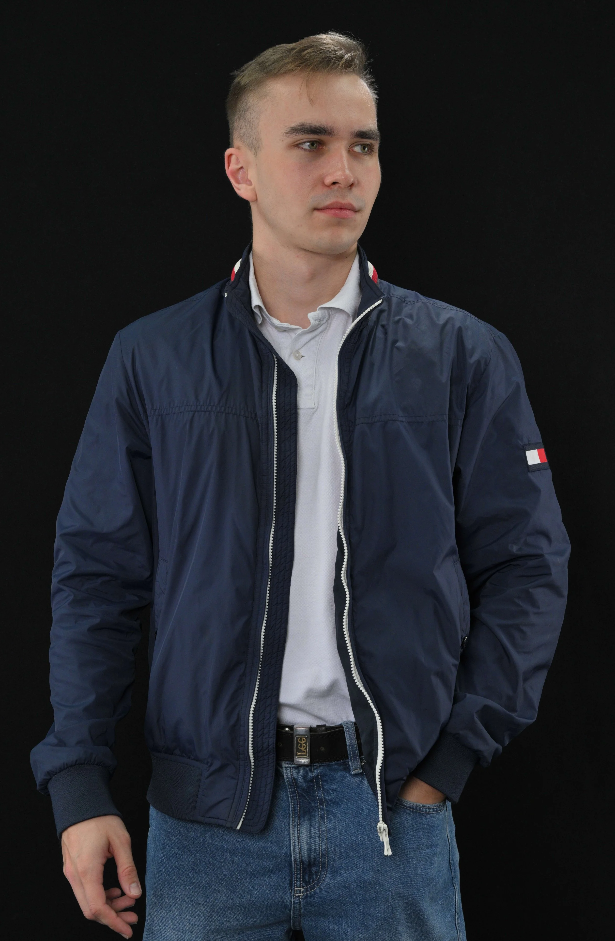 A young man with blonde hair, wearing a navy blue jacket over a white polo shirt and blue jeans, standing against a black background.