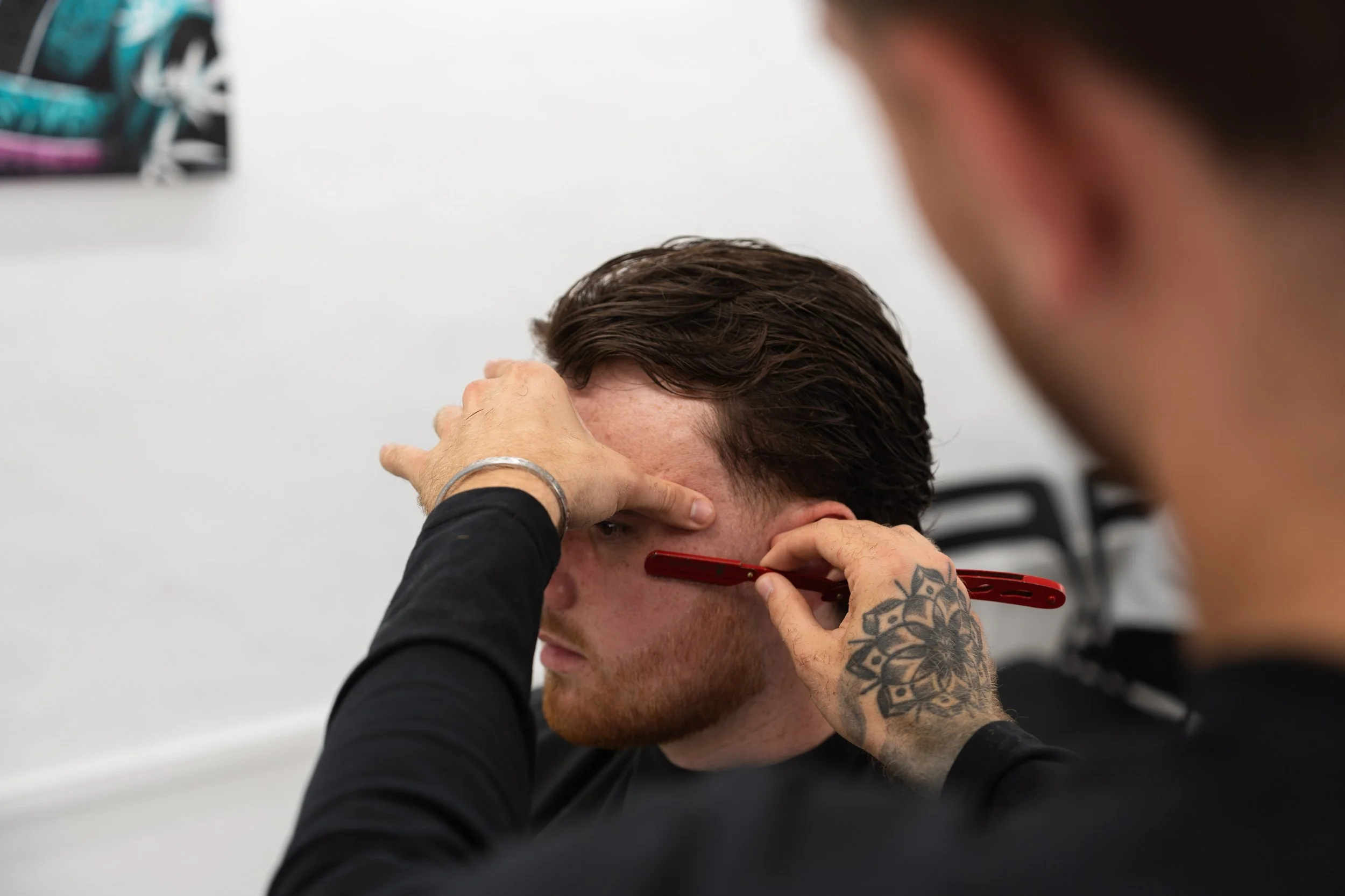 A man with dark hair and a beard is having his face reshaped with makeup, while another person holds a red makeup brush and a hand with a floral tattoo adjusts his facial features.