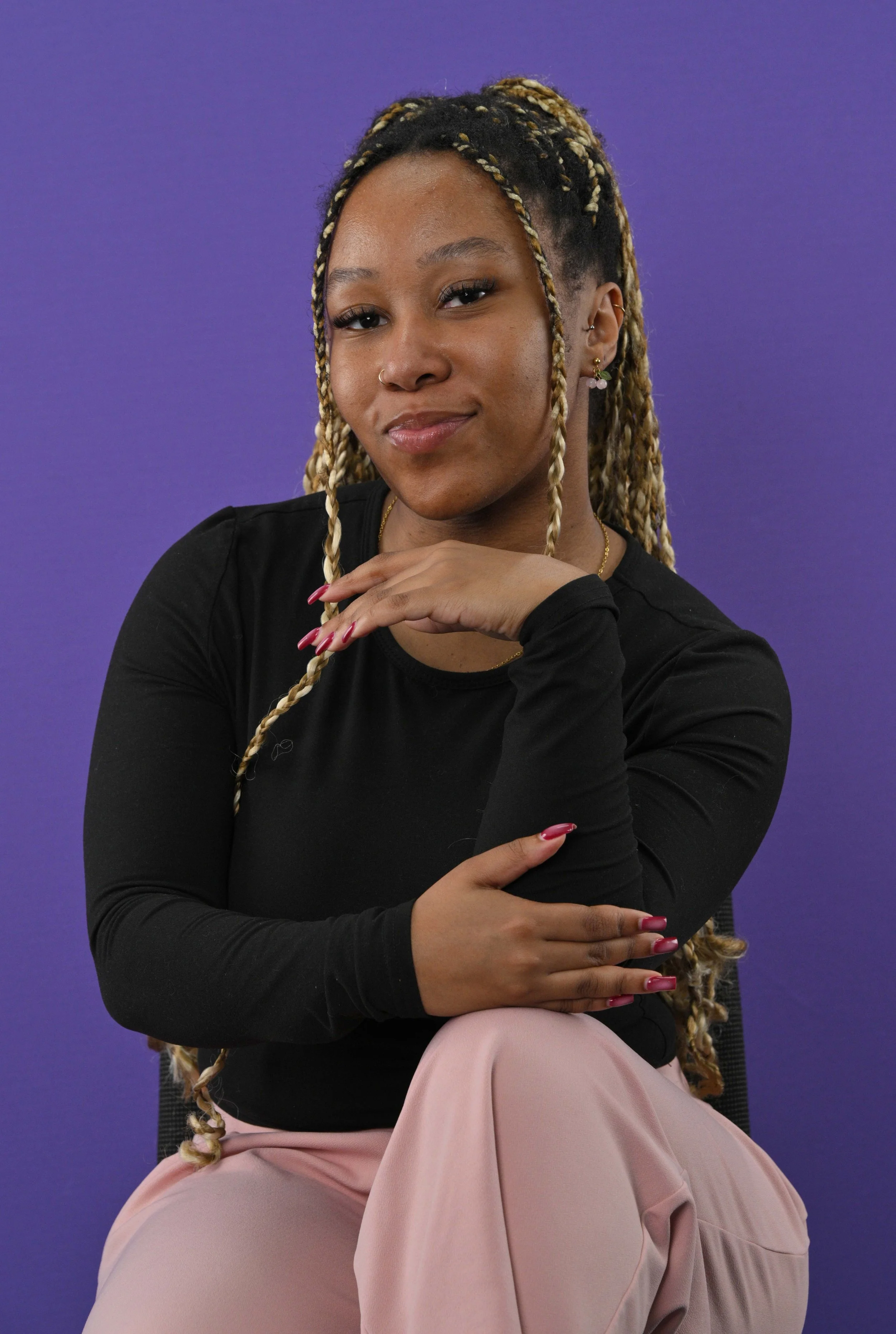 A young woman with blonde braided hair, wearing a black long-sleeve top and pink pants, seated against a purple background, smiling softly at the camera with her hand resting near her chin.