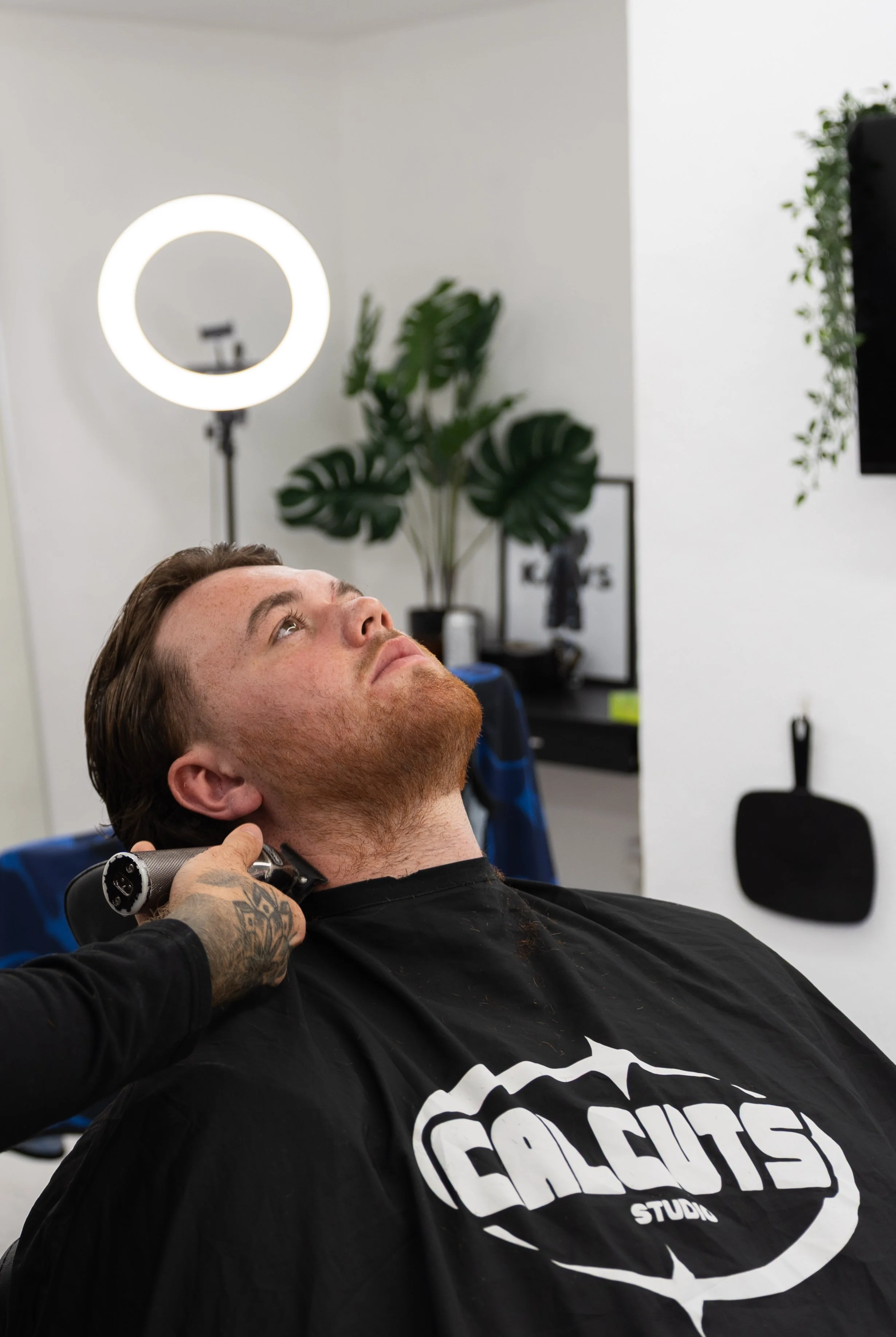 A man with a beard and tattooed hand getting a haircut at a barber's shop. He is seated, leaning back with a black cape that says 'GALSUTÉ STUDIOS,' while a barber trims his hair with clippers. There is a ring light and a green plant in the backgroun
