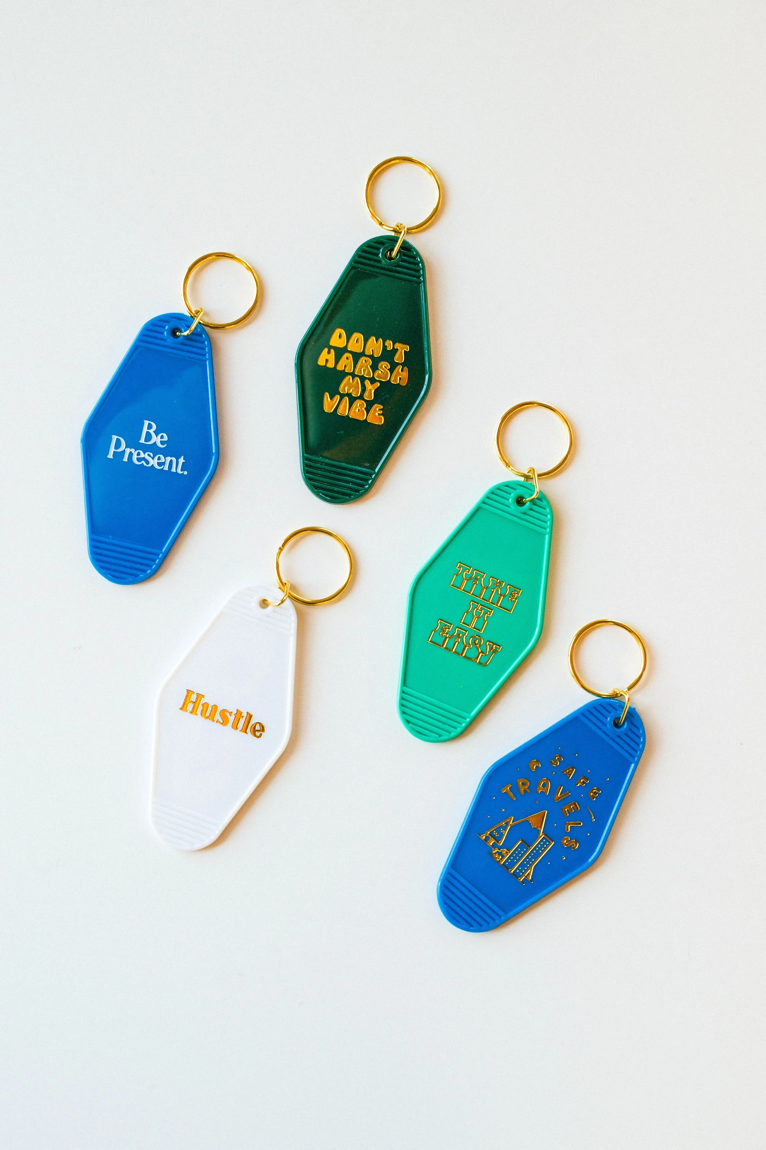 Colorful acrylic keychains with motivational phrases, including 'Be Present,' 'Don't Harsh My Vibe,' 'Hustle,' 'Take It Easy,' and 'Travel Safe,' arranged on a white background.