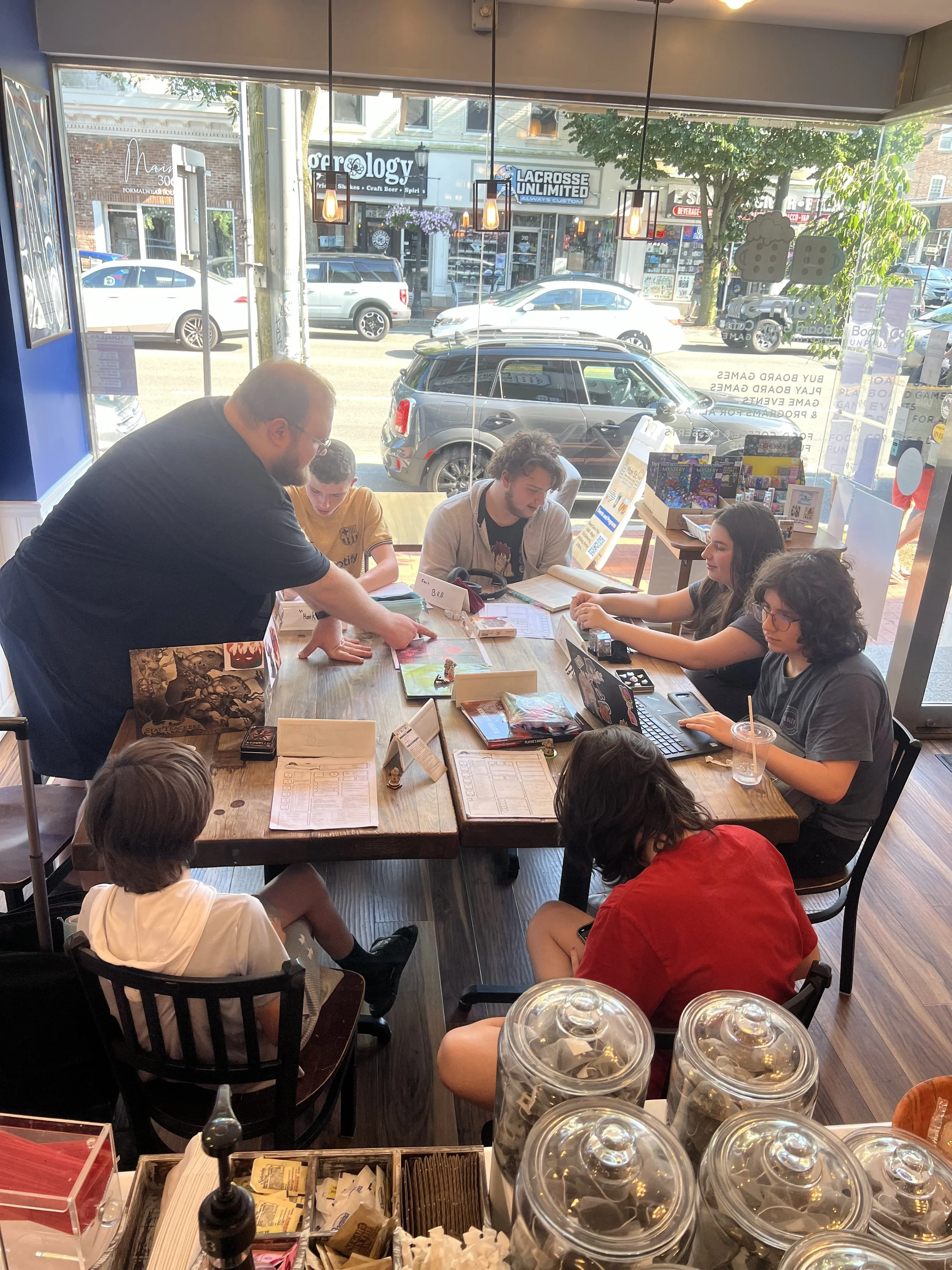 A group of people playing a tabletop role-playing game inside the cafe with large windows facing a street with parked cars and storefronts.