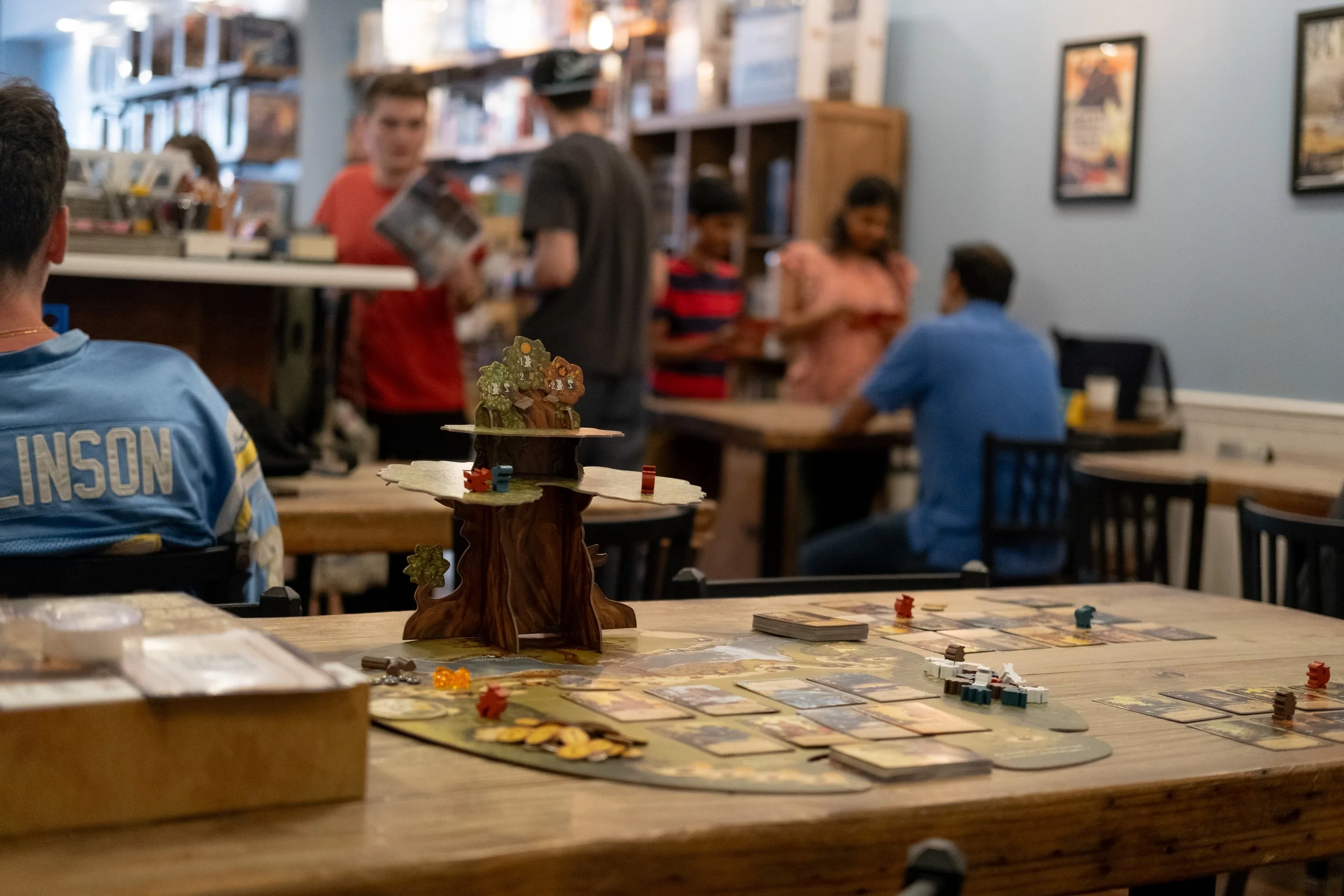 A group of people playing a tabletop board game in a cozy cafe or game store, with a variety of game pieces, cards, and a scenic game tree centerpiece on the table, while others are sitting or standing around in the background.