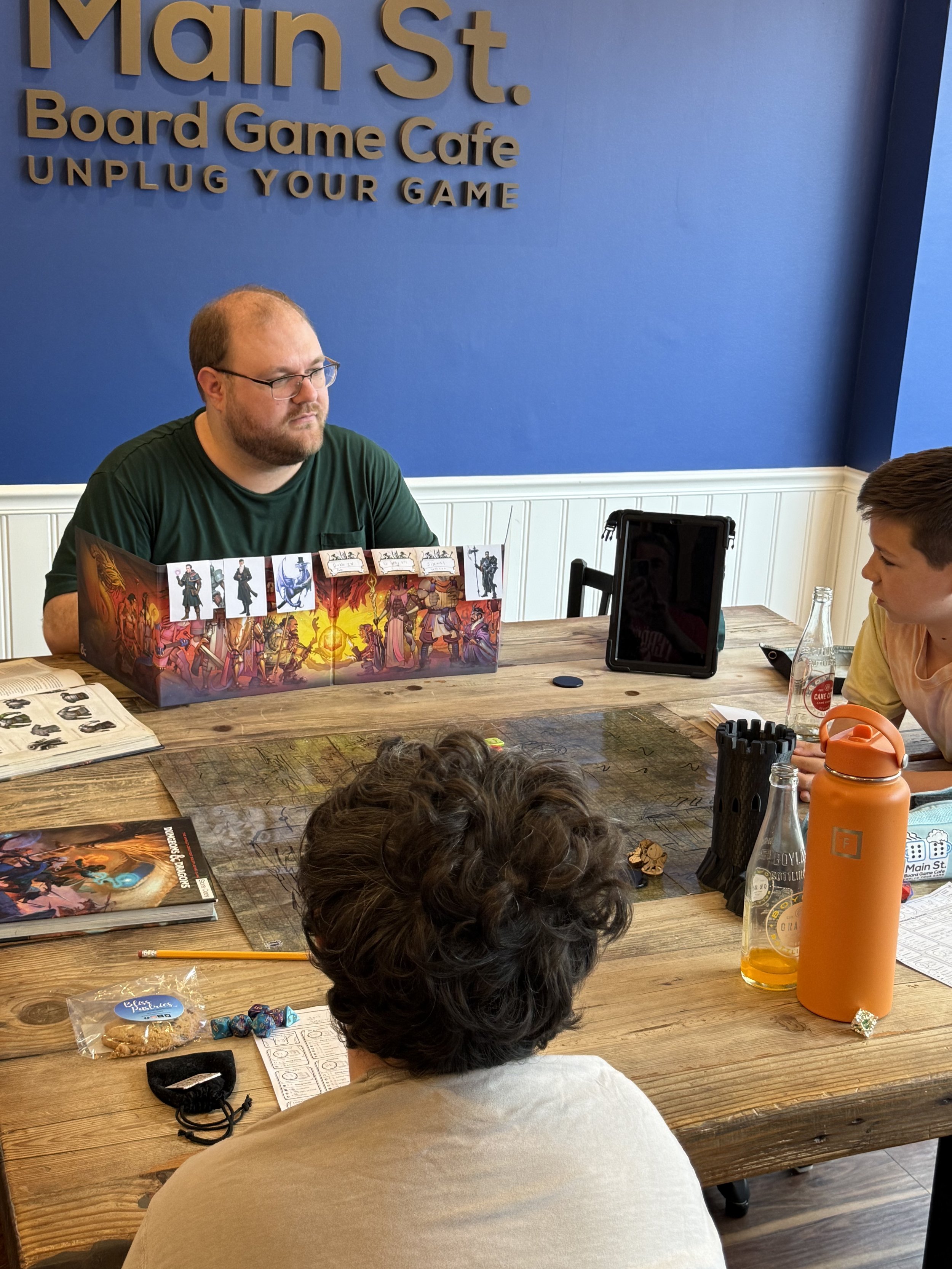 A group of people playing a tabletop board game at the cafe with the game setup, character sheets, dice, and drinks on the table.