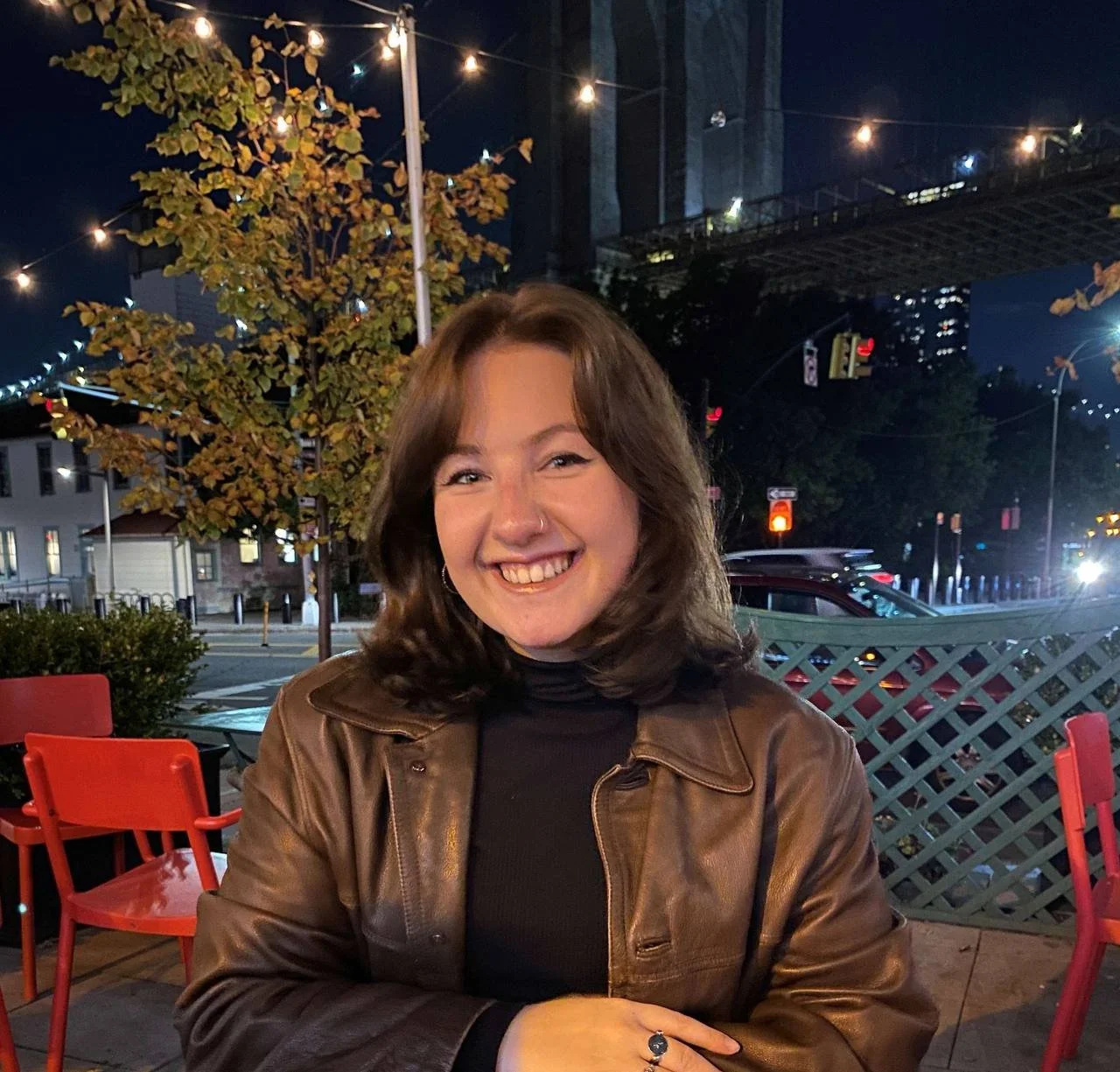 A woman with shoulder-length brown hair, smiling and wearing a brown leather jacket over a black turtleneck, sitting at an outdoor restaurant at night with string lights overhead, a tree, and a cityscape with tall buildings in the background.