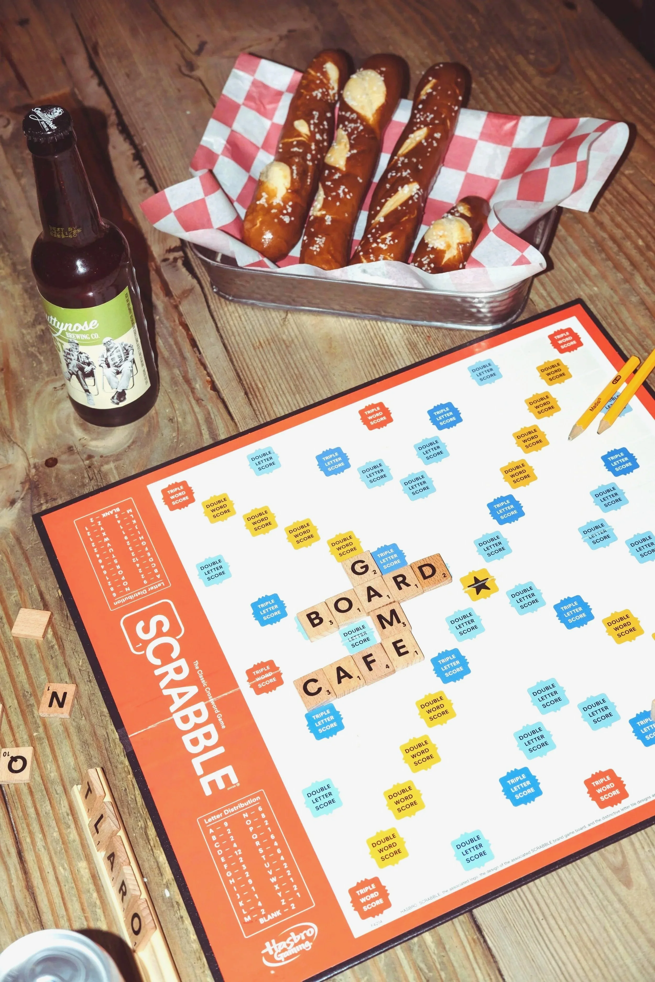 A Scrabble board game with words 'BOARD' and 'CAFE' linked. To the side, tiles spell 'N', 'O', 'L', 'A', 'R', 'O'. A bottle of beer and a basket of churros are on the wooden table.