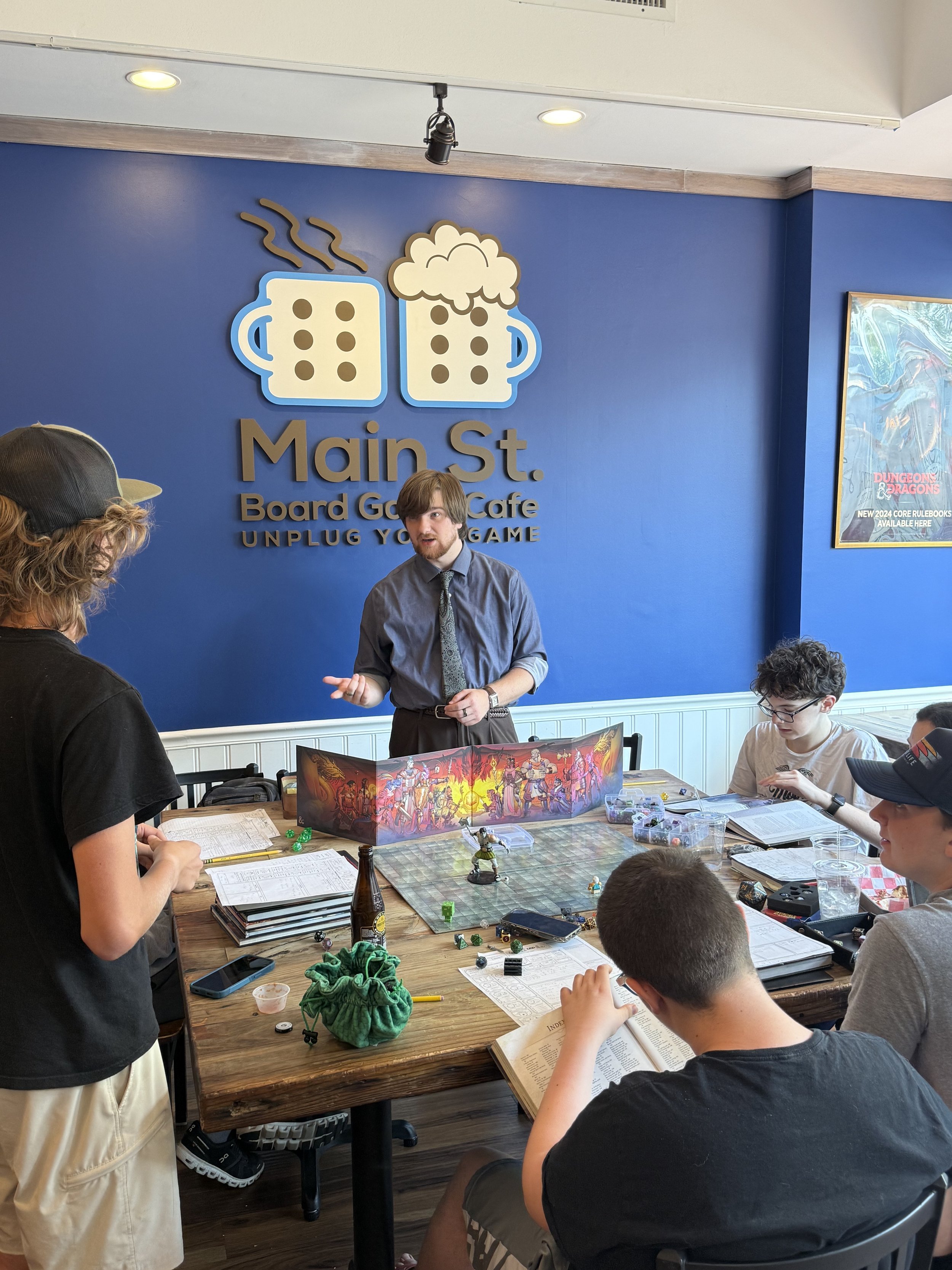 A group of people playing Dungeons & Dragons inside a cafe with a blue wall and a sign that reads 'Main St. Board Game Cafe.' The sign features two coffee mugs, one steaming and one topped with foam. A man stands at the head of the table explaining the game while others sit around the table with game materials, books, and dice.