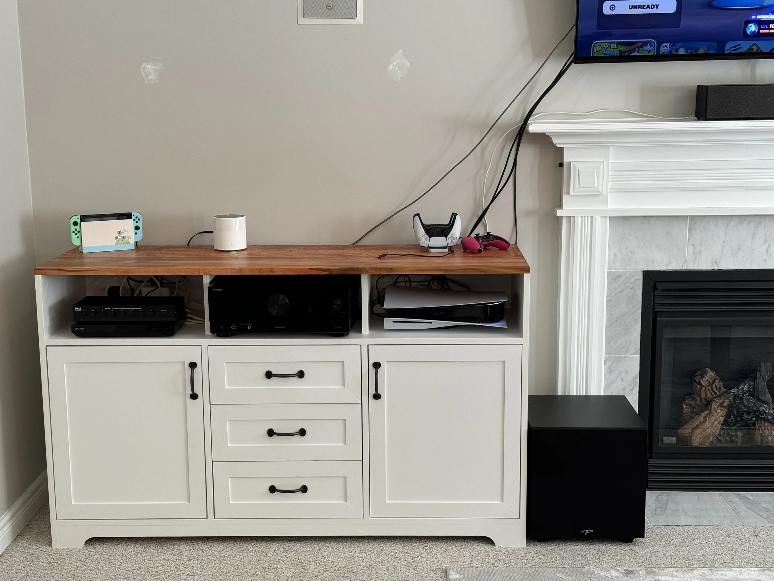 Living room entertainment area with a white cabinet holding gaming consoles, a white smart speaker, and a controller. There is a TV mounted on the wall. A small dog is sitting on the carpet near the black speaker and below the fireplace.