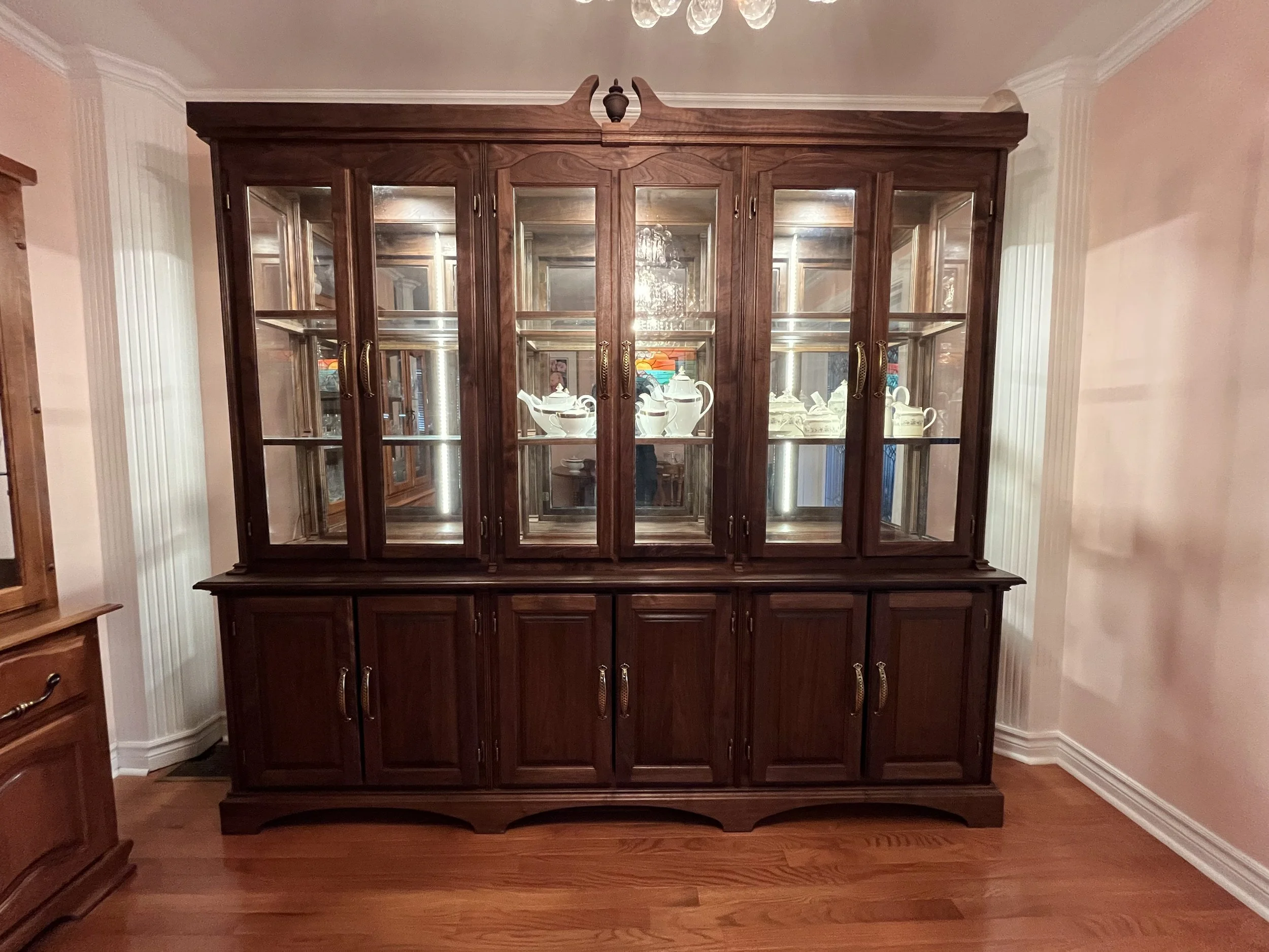 A large wooden display cabinet with glass doors, filled with white teapots, tea sets, and decorative items, positioned against a pink wall with crown molding.