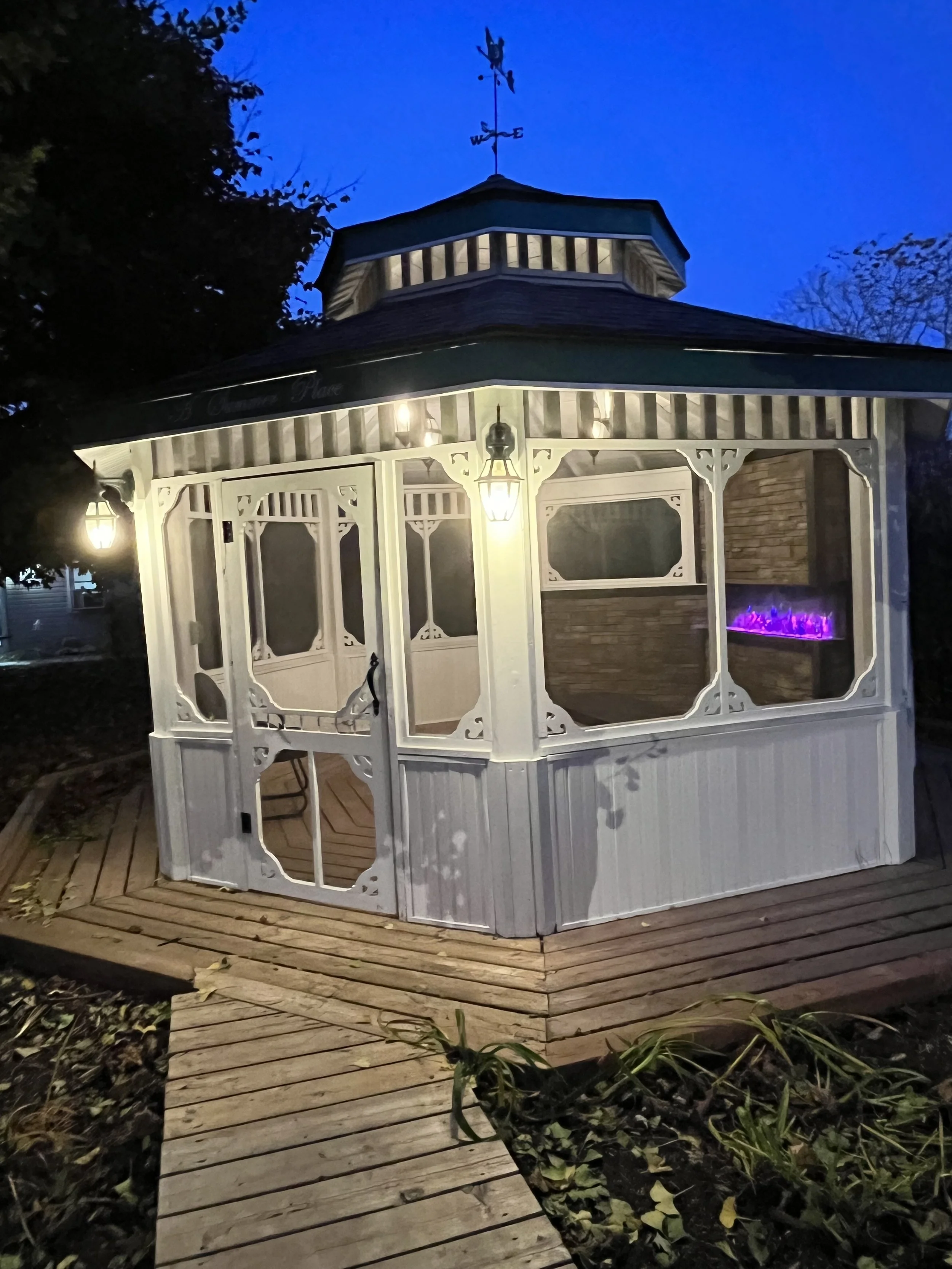 A small, white, covered outdoor pavilion with decorative trim, illuminated by hanging lanterns, situated on a wooden deck with a pathway leading up to it, surrounded by garden plants, during dusk or early evening.