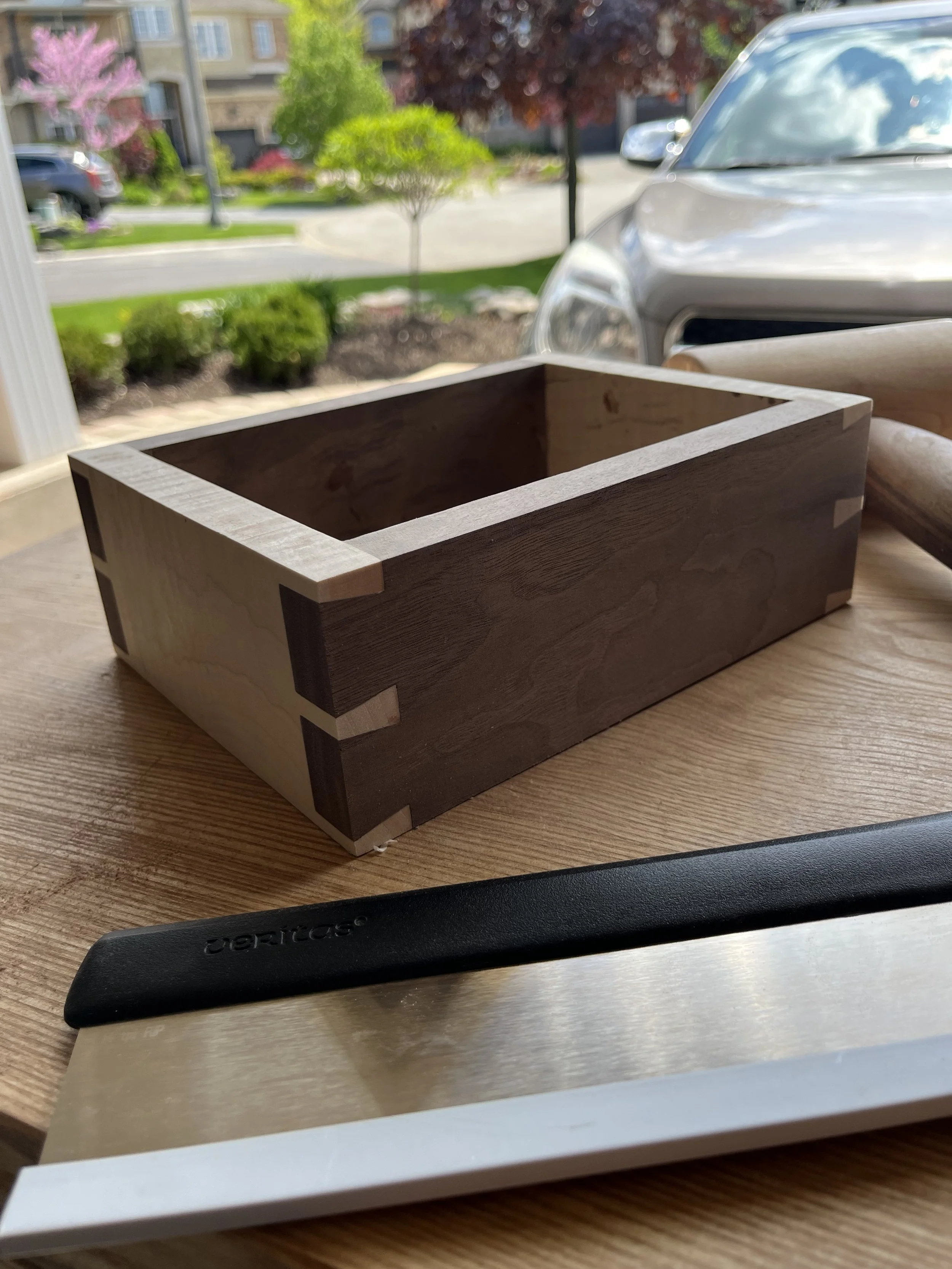 Close-up image of a wooden box, a chef's knife with a black handle and stainless steel blade, and a metal ruler on a wooden surface.