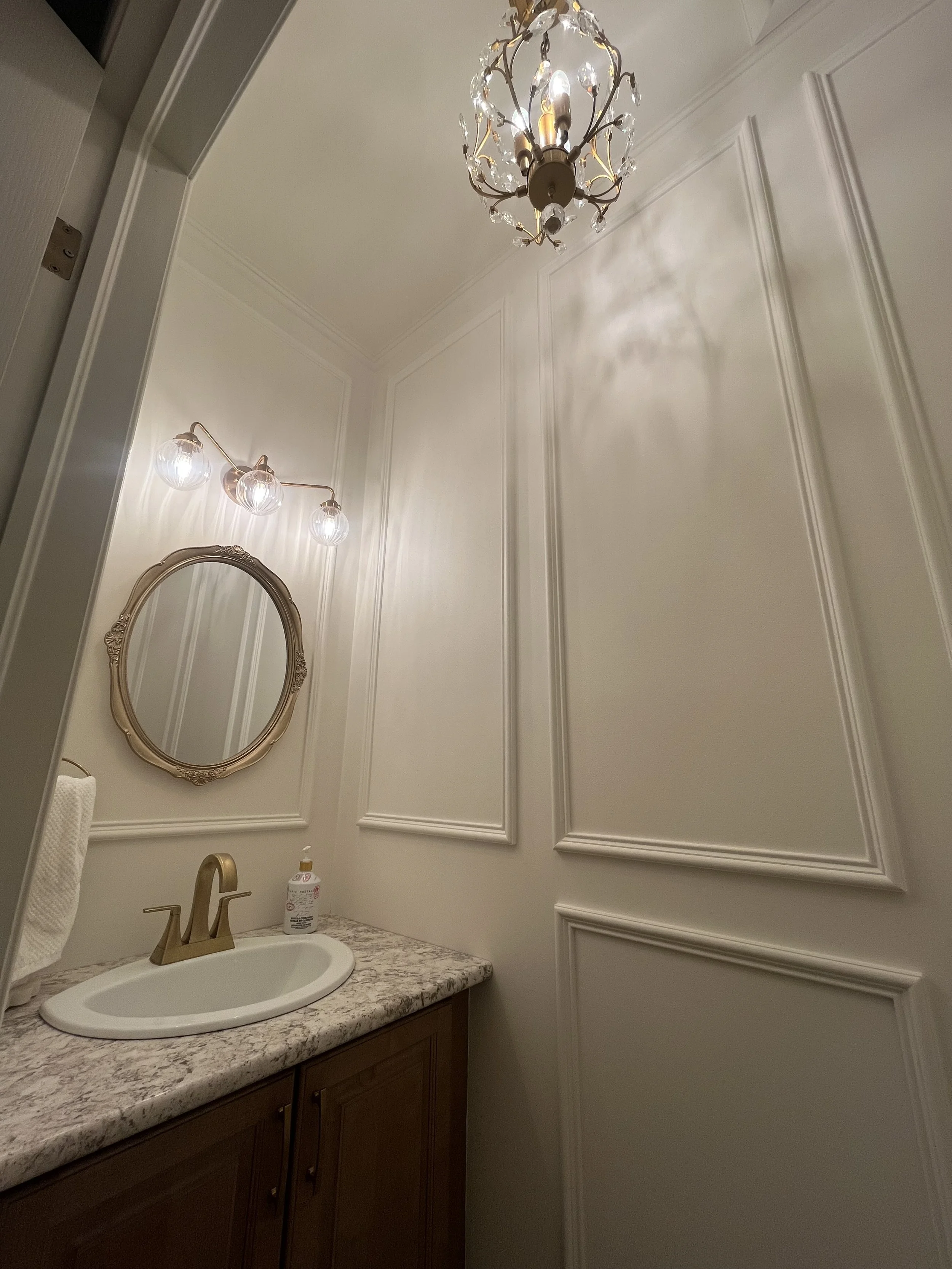 A bathroom with white walls featuring decorative molding, a marble countertop with a sink, a gold faucet, and a mirror. The bathroom has a chandelier with crystal accents and a wall-mounted light fixture with three glass globes. A hand soap dispenser is on the counter.