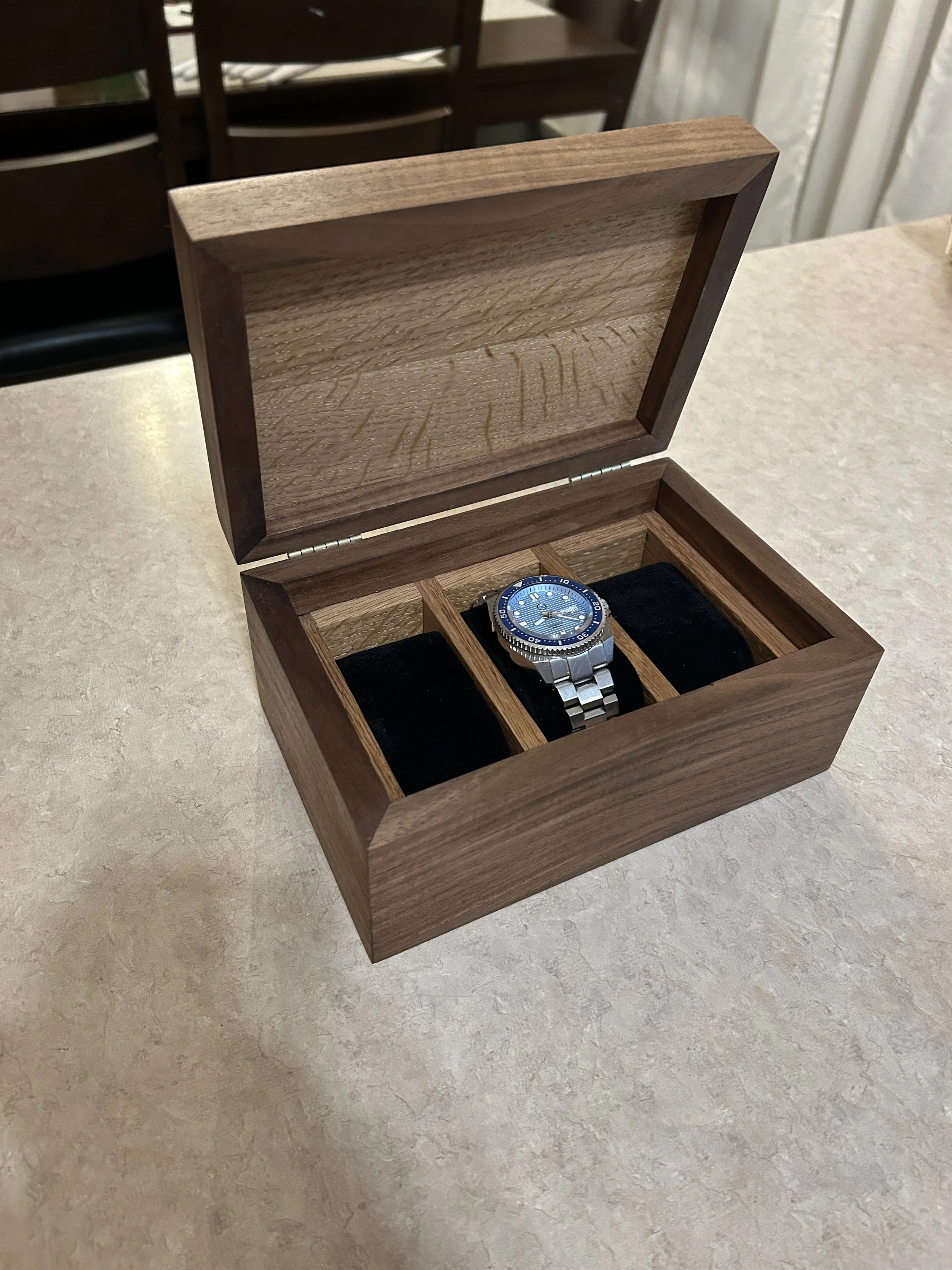 A wooden watch box with a hinged lid, containing a blue wristwatch on a black cushion in one of its compartments. The box is placed on a light-colored surface, with dining chairs and curtains visible in the background.