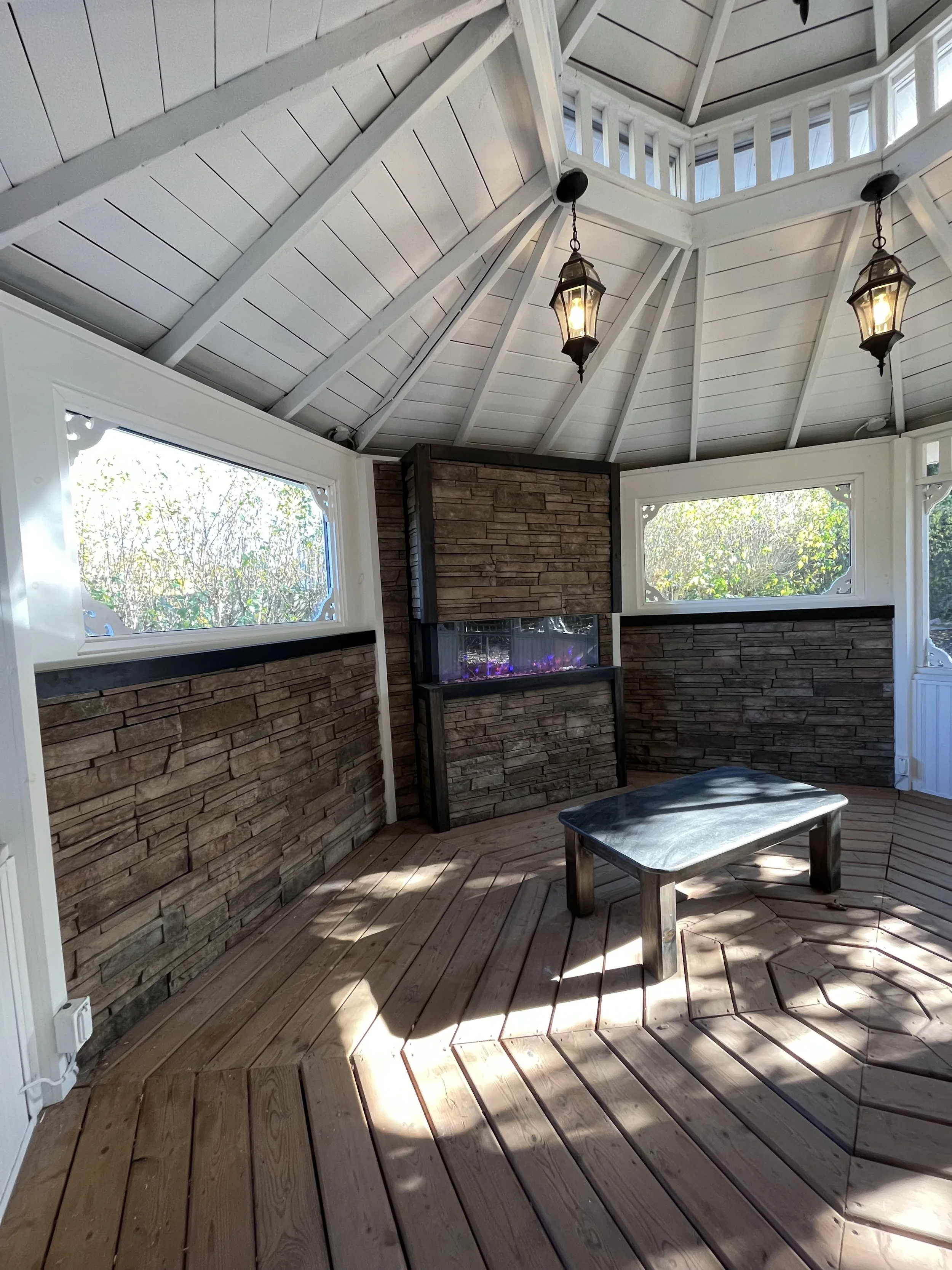 A room with a high, white, wooden ceiling with two hanging lantern-style lights. The walls are covered with stone veneer, and there are large windows letting in natural light. There is a small wooden table on the wooden floor.