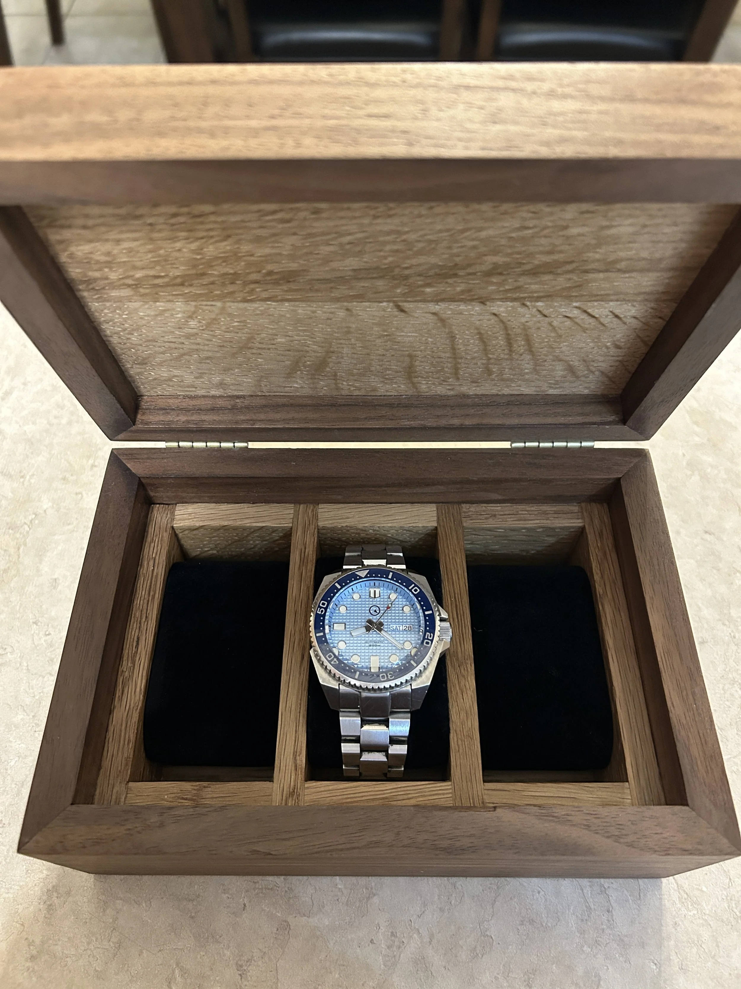 A silver wristwatch with a blue face in a wooden watch box with multiple compartments.