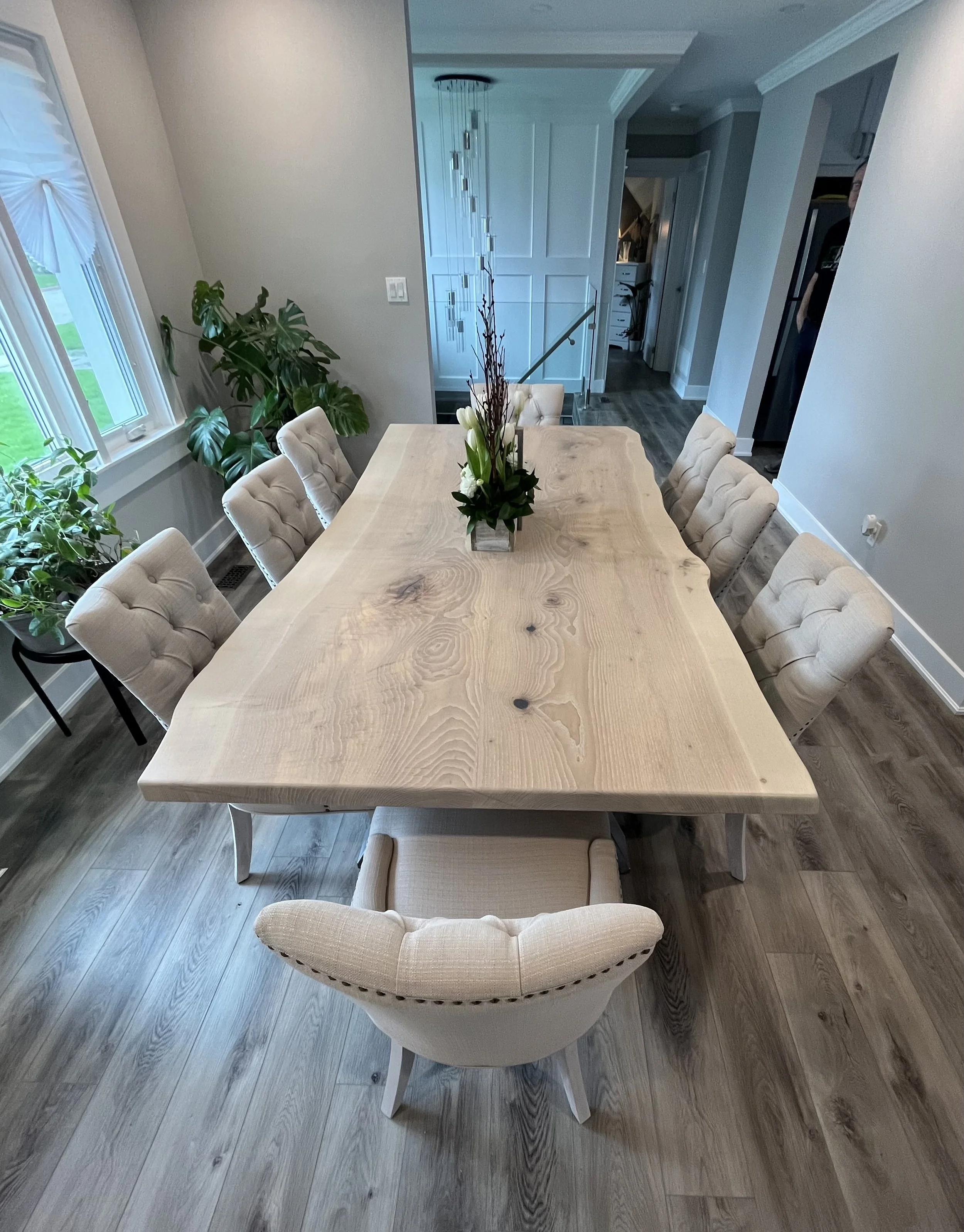 A dining room with a large wooden table surrounded by upholstered beige chairs. A floral centerpiece is on the table. There are large windows with white and blue curtains on the left, and a potted plant in the corner. The room has wood flooring, crown molding, and a staircase in the background leading to an upper level.