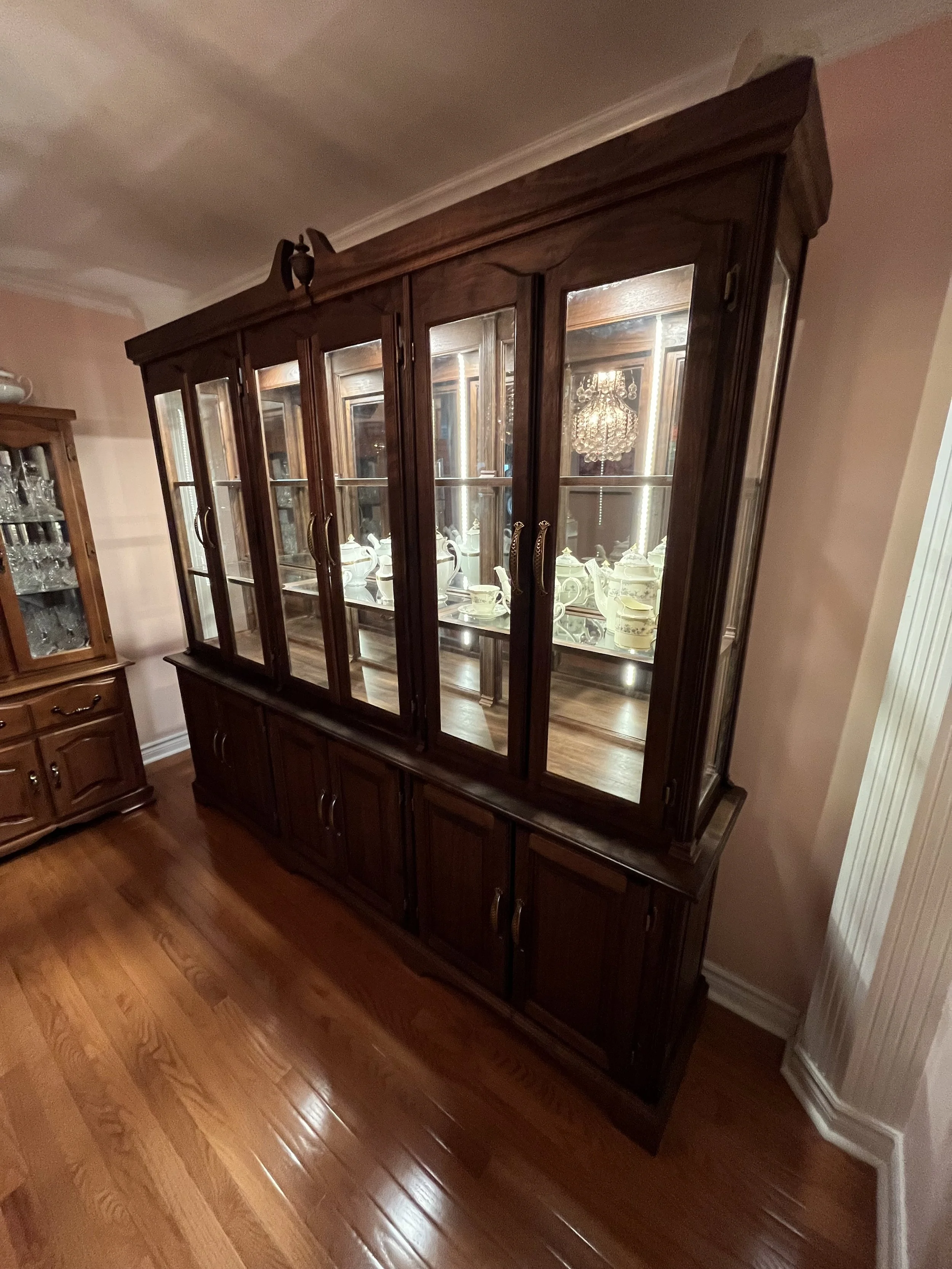 A large wooden china cabinet with glass doors displaying fine china and glassware, in a dining room with hardwood floors and pink walls.