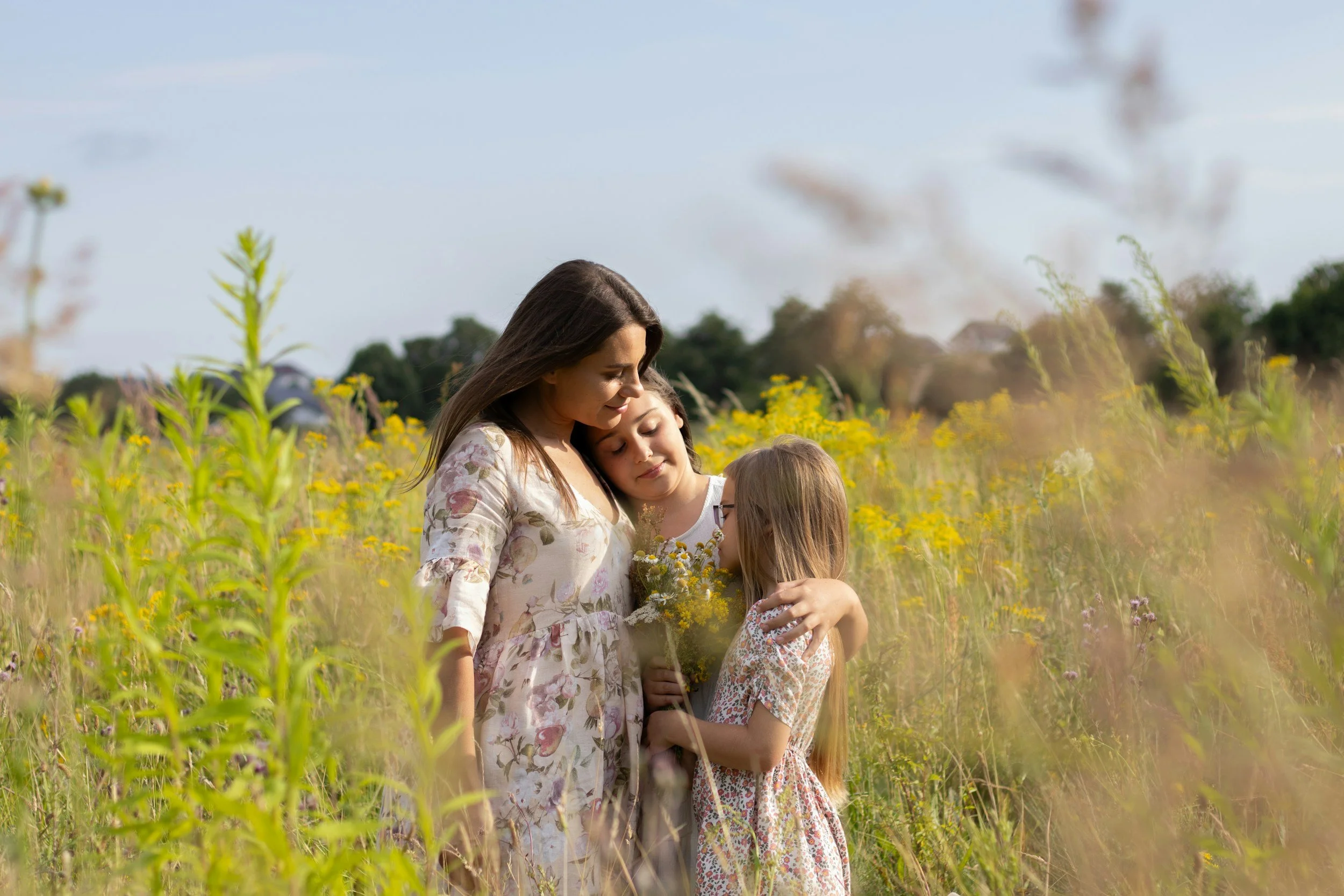 A woman and two young girls in a field of yellow flowers, sharing a tender moment, with a clear sky and trees in the background.
