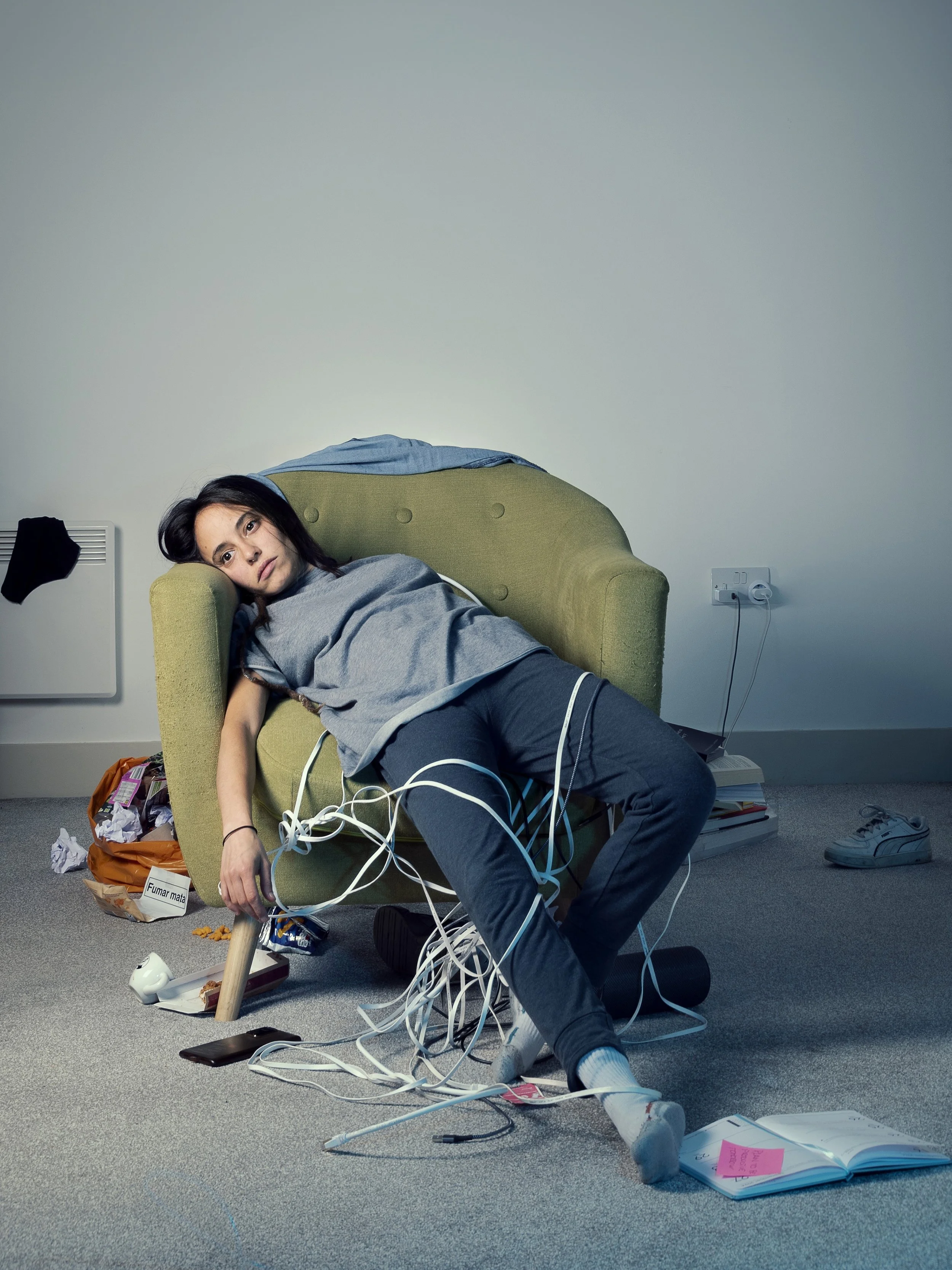 A woman is sitting on the floor, slumped against a green armchair with tangled cords and electronic devices around her. She appears exhausted or distressed with cluttered surroundings and scattered items.