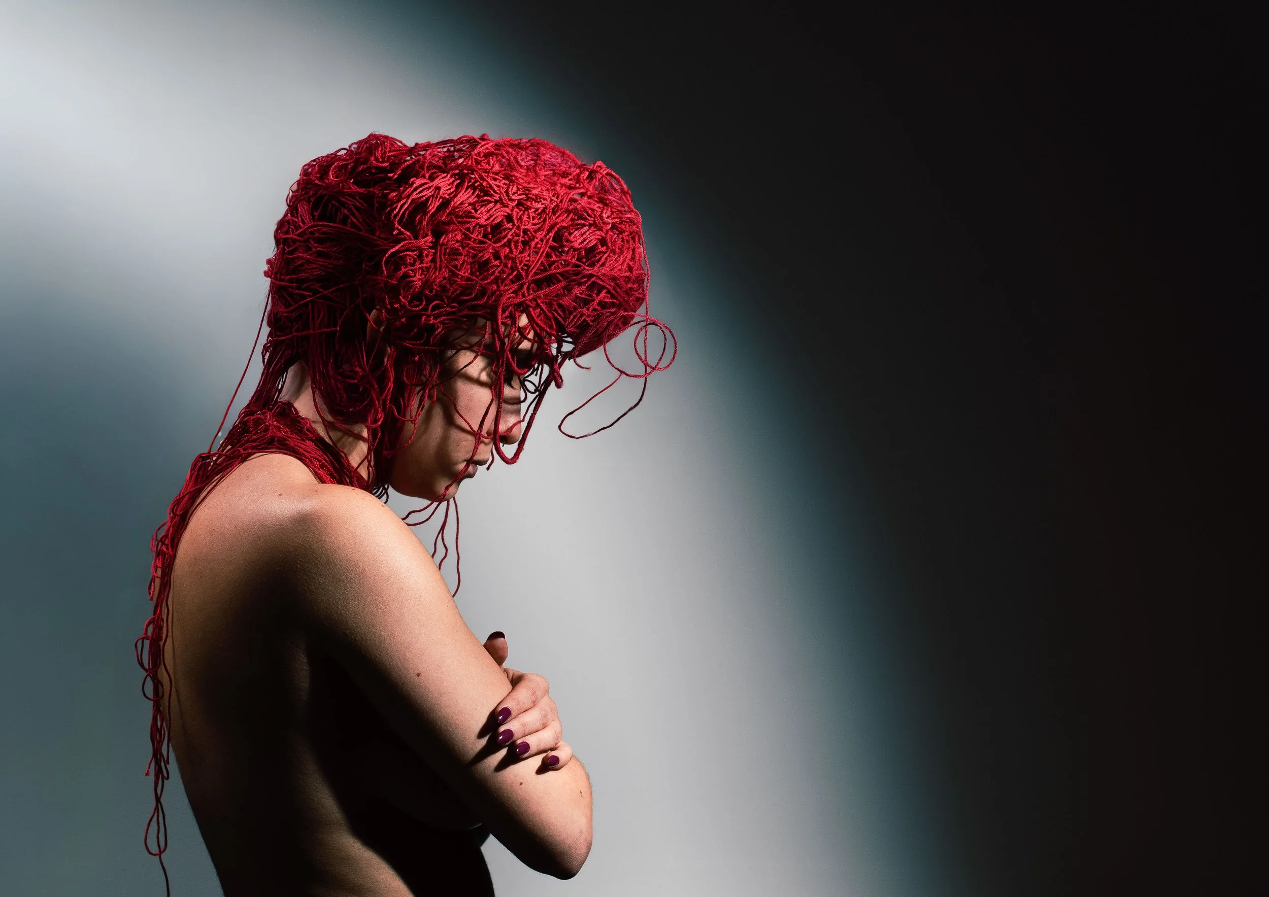 A woman with red yarn intertwined in her hair, standing topless with her arms crossed, against a gradient background.