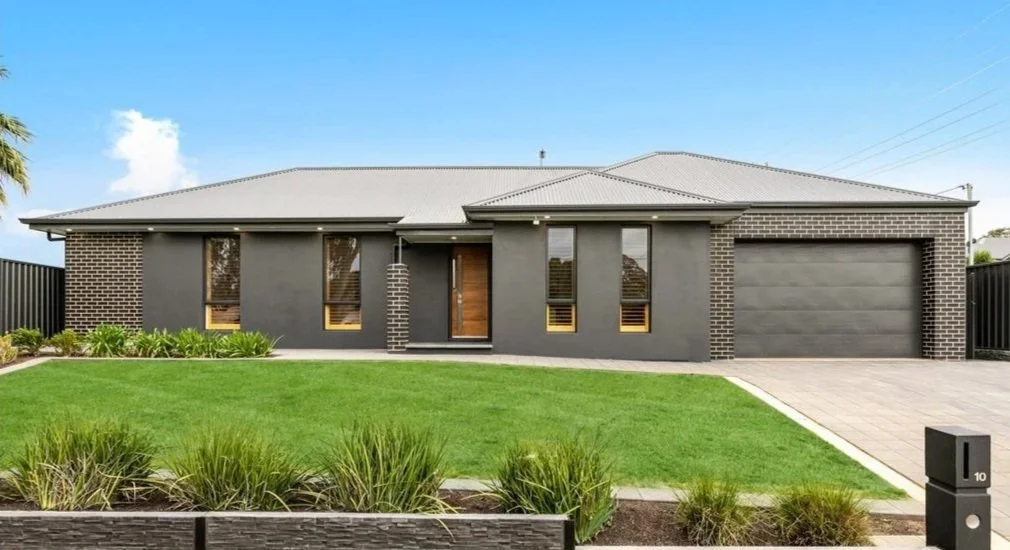 Modern single-story house with a gray exterior, brick accents, and a landscaped front yard with green grass and plants, a paved driveway, and a garage.