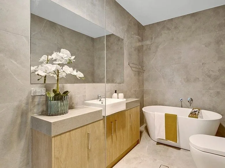 Modern bathroom with gray tile walls, a large mirror, a white rectangular sink on a wooden cabinet, a vase with white flowers, a bathtub with a yellow towel, and a small shelf.