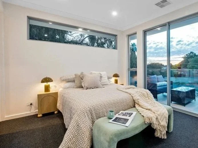 Modern bedroom with a bed covered in white patterned bedding and multiple pillows, two bedside tables with yellow lamps, a light green cushioned bench with magazines and a throw blanket, large sliding glass doors opening to a balcony with outdoor sea