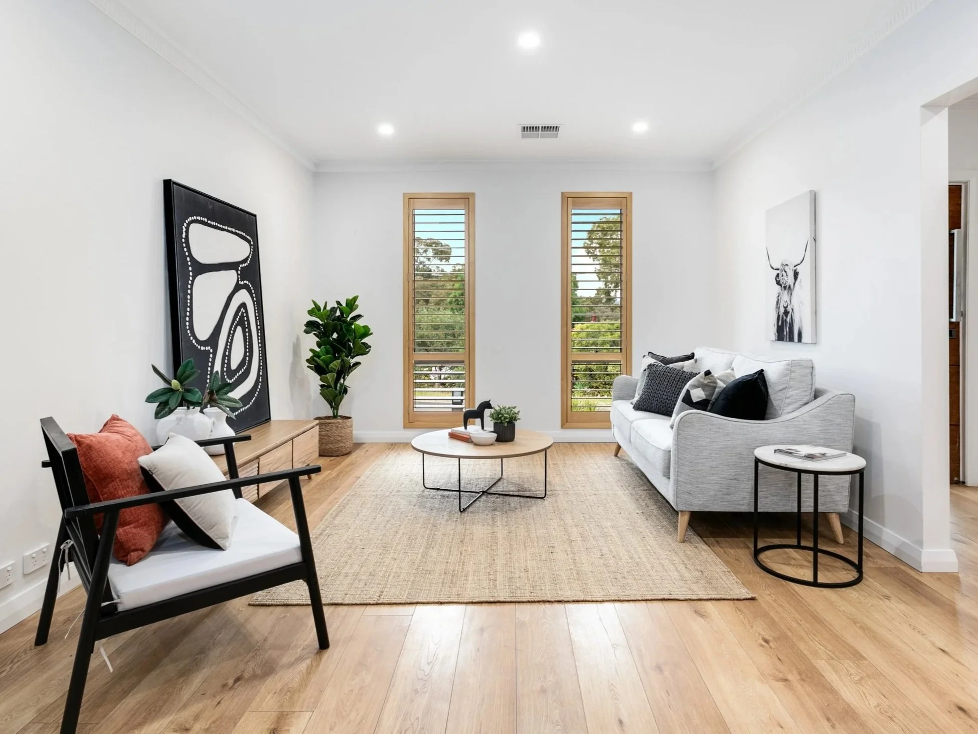 Modern living room with white walls, light wood flooring, a white sofa with black and gray pillows, two tall windows with wooden shutters, black and white artwork, a black armchair with pillows, a round coffee table, a side table, a large potted plan