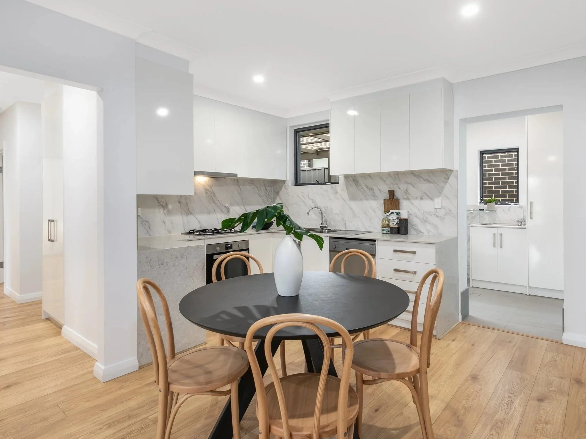 Modern kitchen with white cabinets, marble backsplash, black round dining table with wooden chairs, and a vase with green leaves.