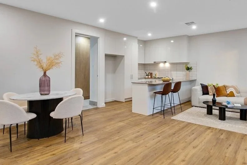 Living room and kitchen area with hardwood flooring, white walls, a round dining table with white chairs, a large vase with dried flowers, a beige sofa with pillows, a black coffee table, and a white kitchen with bar stools.