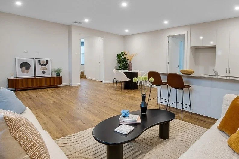 Modern living room with white walls, hardwood floors, a black coffee table, and a beige sofa with throw pillows. There is a kitchen island with barstools and minimal decor.