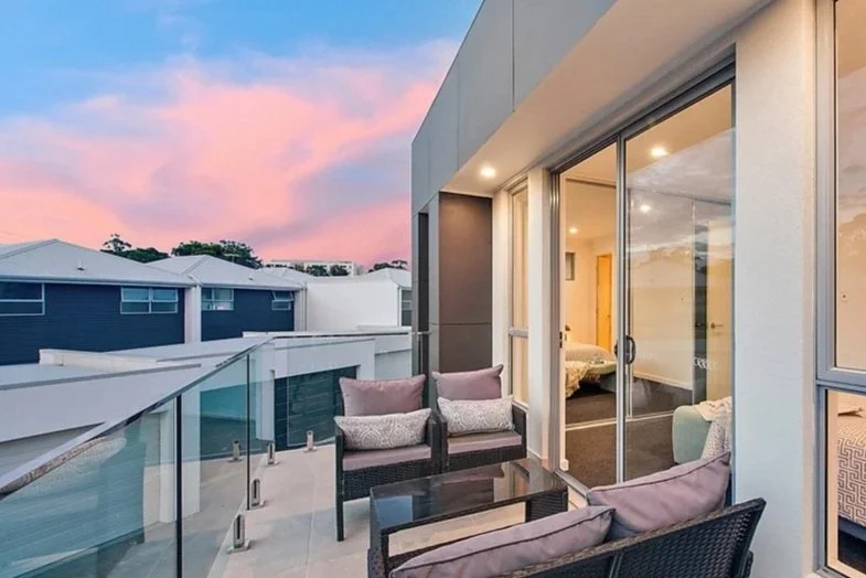 Balcony with outdoor seating including two chairs and a sofa with cushions, glass railing, view of neighboring houses, sunset sky, and interior bedroom visible through sliding glass doors.