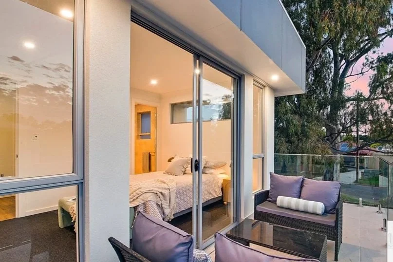 Modern bedroom visible through sliding glass doors, with outdoor seating on a balcony, trees in the background