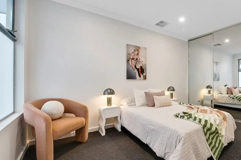 Modern bedroom with a bed, nightstands, lamps, an armchair by the window, and a large mirror closet in the background.