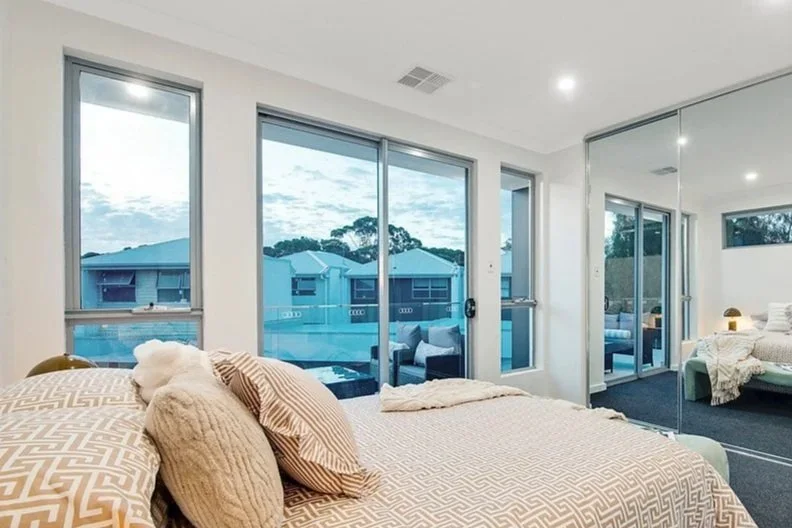 Modern bedroom with large windows and sliding glass doors, mirror wardrobe, bed with beige and patterned pillows, across from a seating area outside, in a well-lit room.