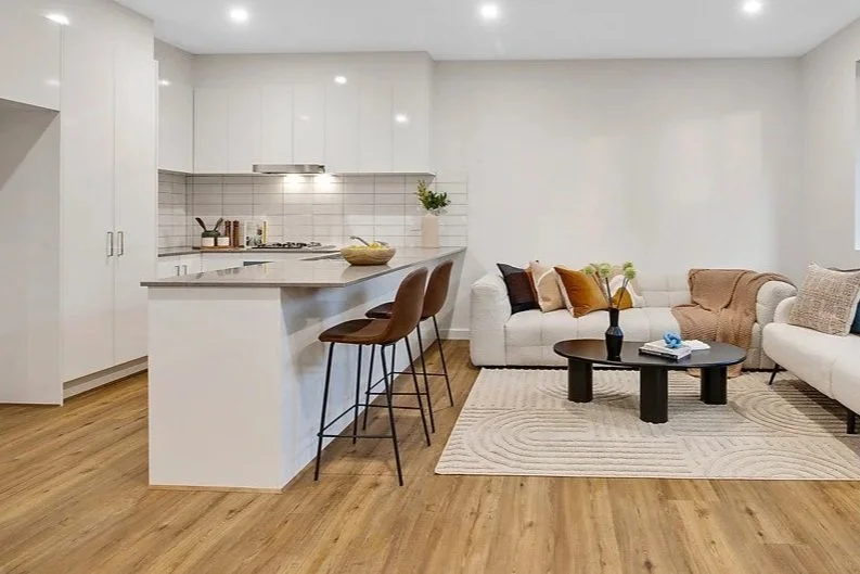 Modern open-plan living room and kitchen with white cabinets, wooden flooring, and neutral furniture. The kitchen has a breakfast bar with three brown chairs. The living area features a white sofa with decorative pillows, a round black coffee table, 